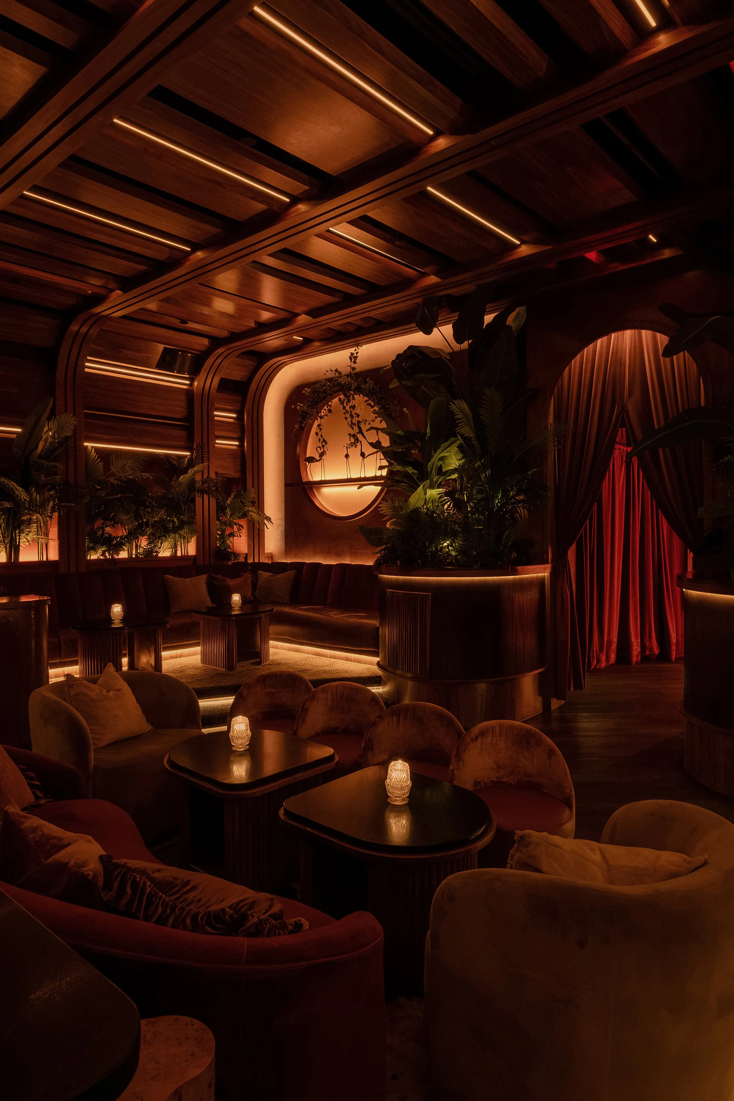 Moody lounge seating at Powder Room with velvet chairs, candlelit tables, dark wood finishes and a dramatic red curtain.