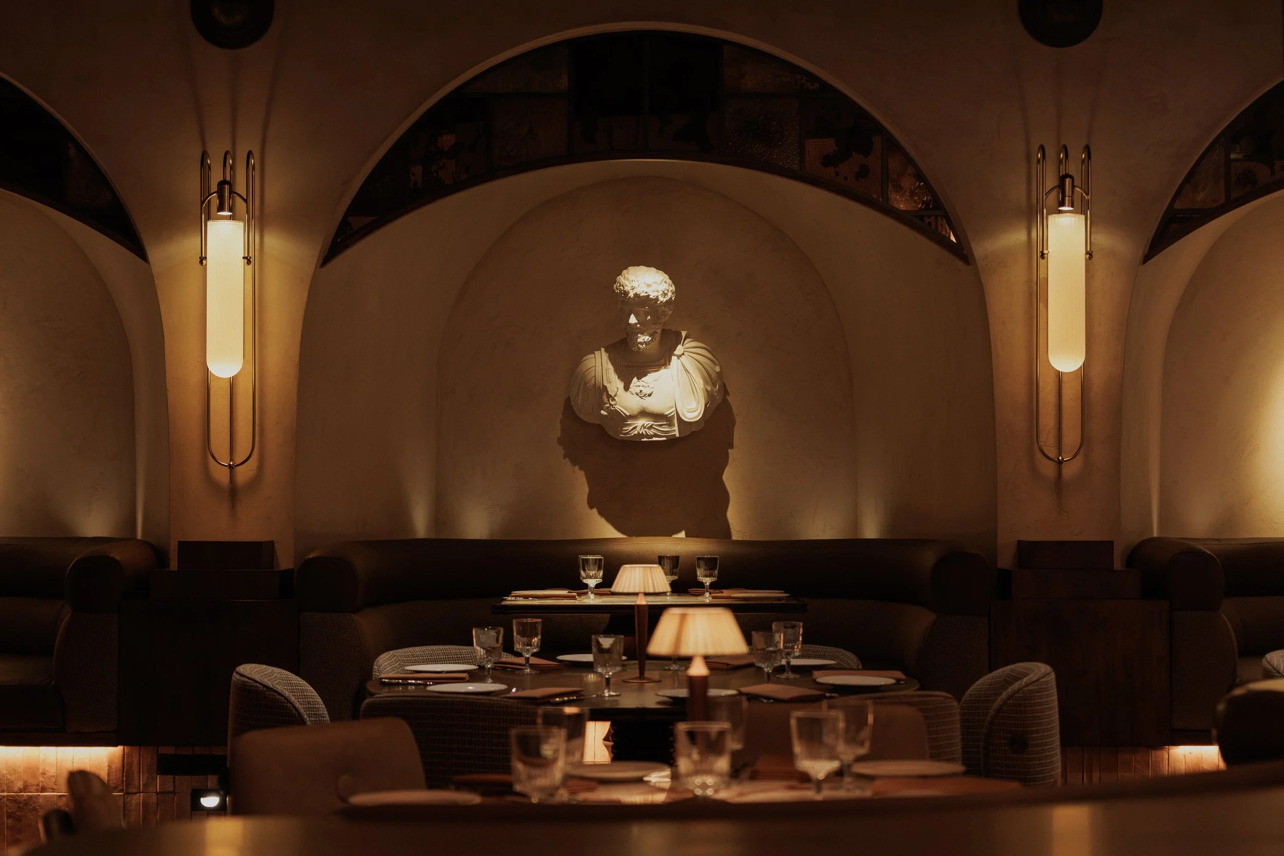 Arched dining alcove at Roman God of Fire with a classical bust, sculptural wall lighting, curved seating and candlelit tables.