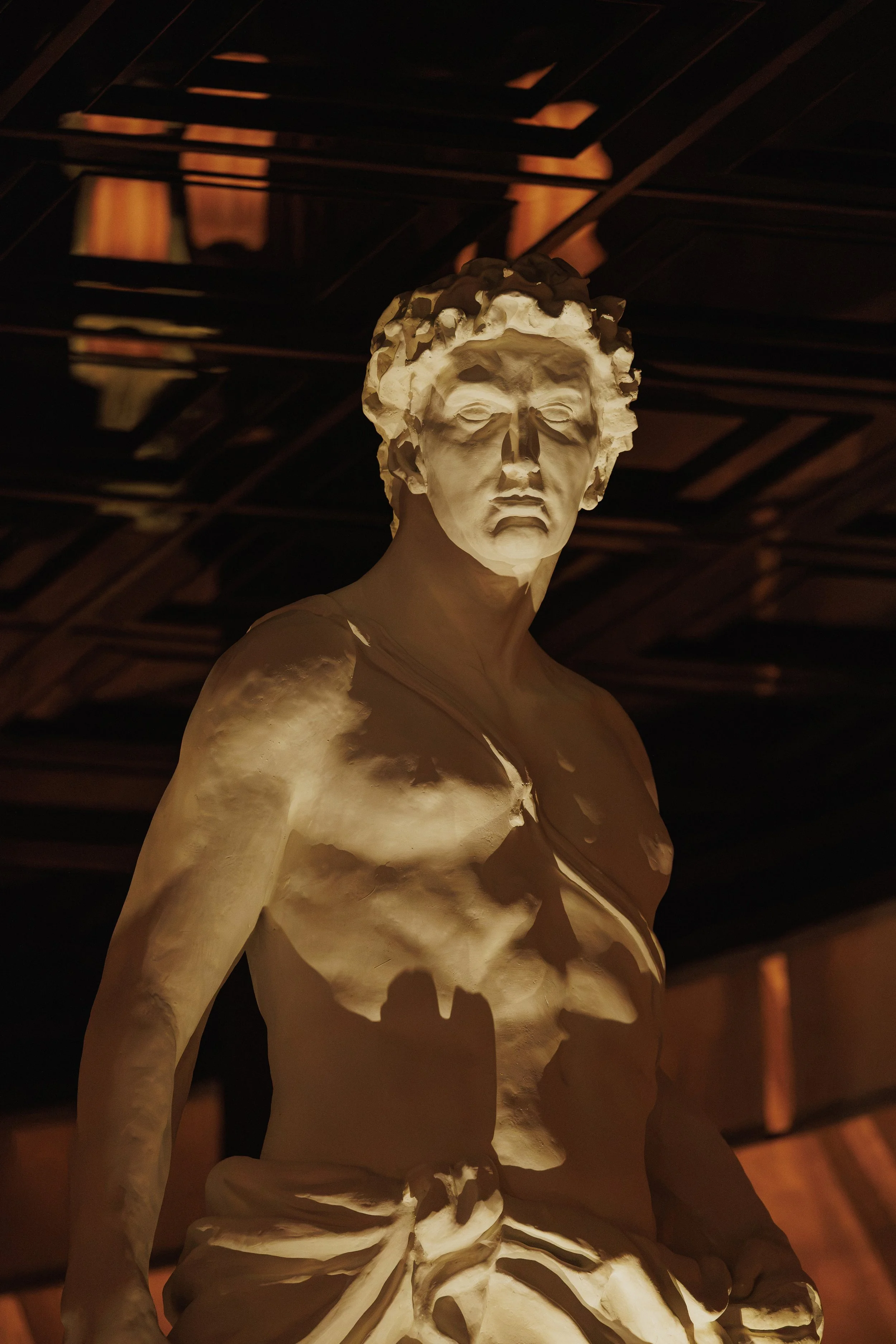 Dramatically lit classical statue of the god Apollo rising beneath the dark geometric ceiling at Roman God of Fire.
