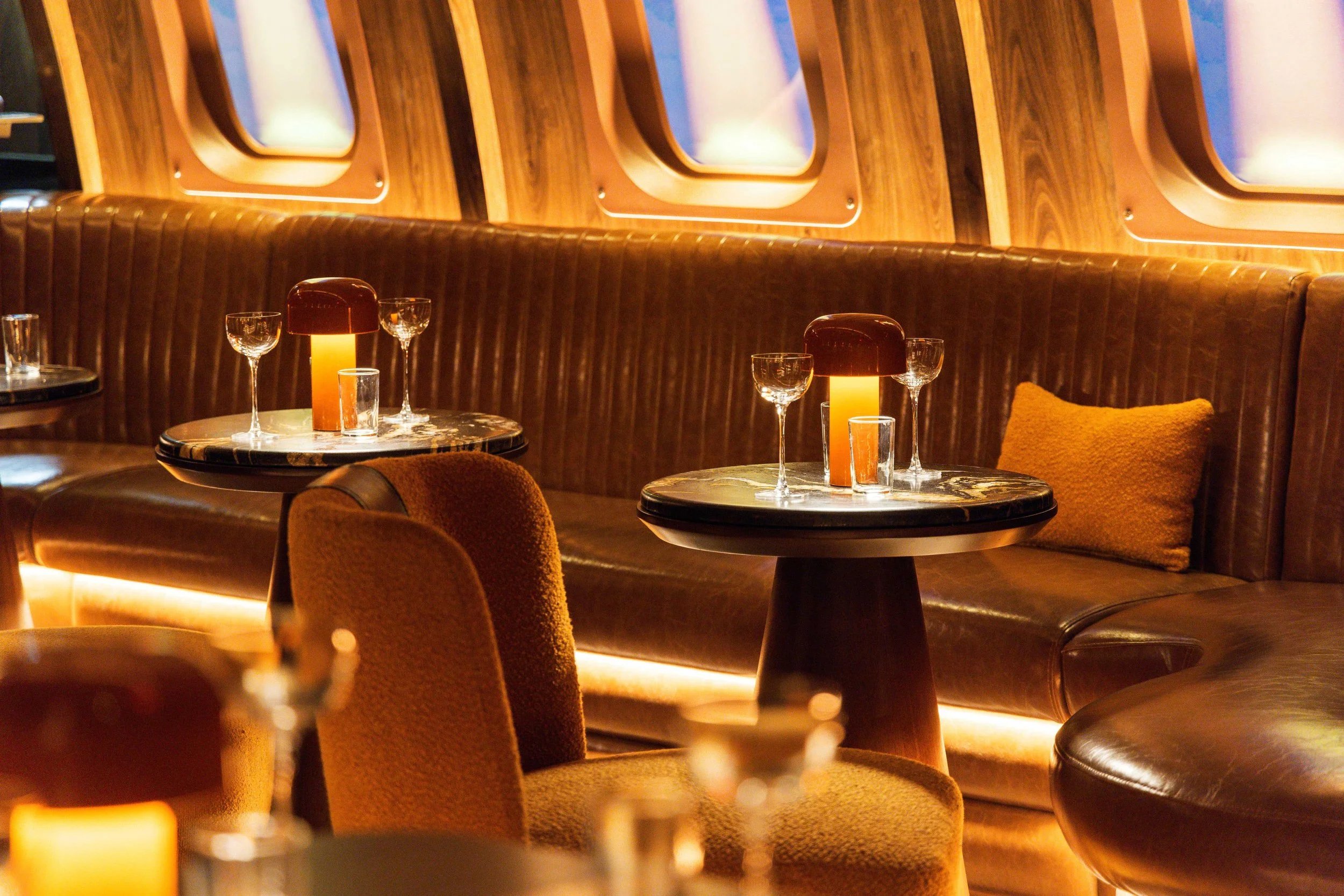 Carry On cocktail bar in Phoenix, designed by Peter Bowden and PETERMAX Co., featuring a vintage airline-cabin interior with wood-paneled walls, airplane-style windows, leather banquette seating, warm amber lighting, and small cocktail tables.