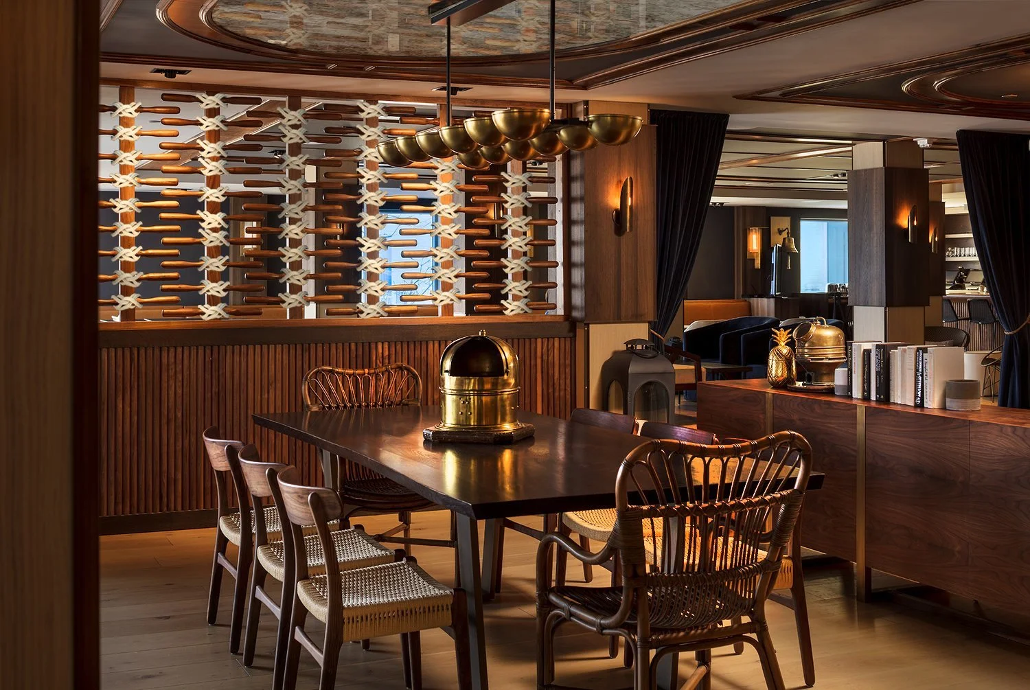 Interior of a stylish restaurant with a dark wooden table, several chairs, a golden dome-shaped object, a bookshelf, decorative items including a pineapple, a vintage vacuum, and a gold pineapple ornament. The background features a partition with a m