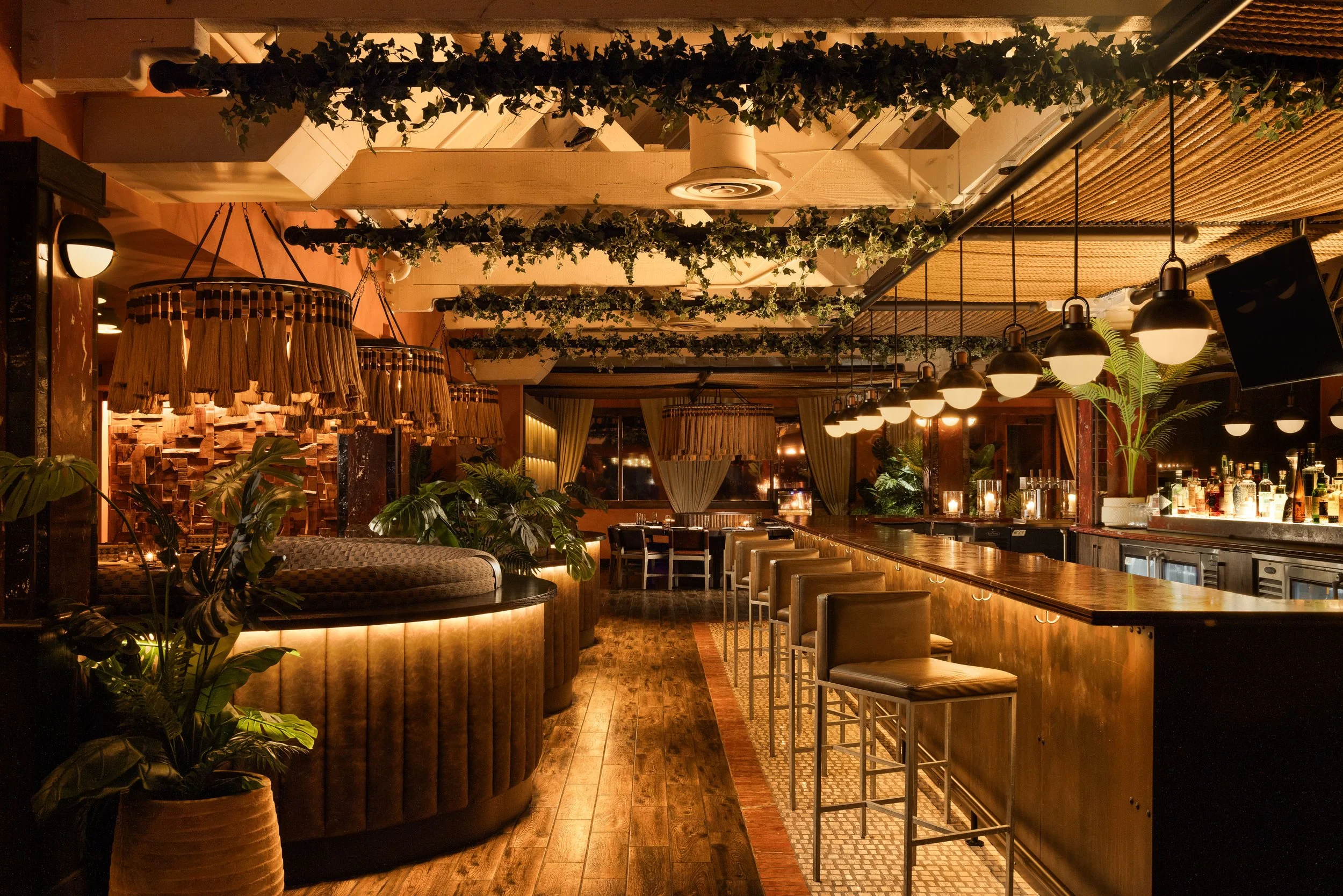 Curved upholstered banquette inside the redesigned Toro Scottsdale, with a green stone table, warm pendant lighting, tropical plants and the restaurant’s bar beyond.