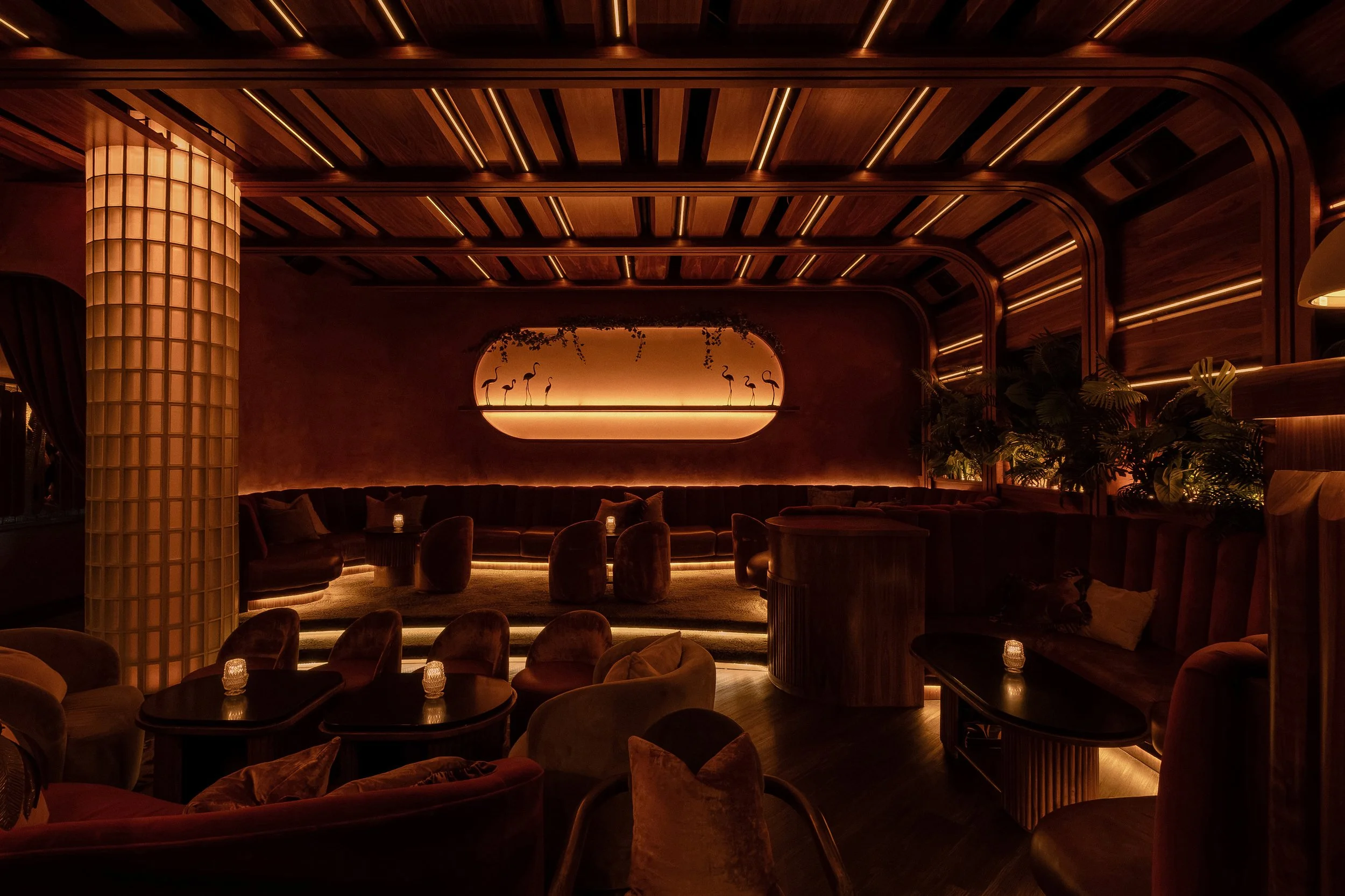 Plush cocktail seating at Powder Room beneath a paneled wood ceiling and oversized glowing wall fixture.