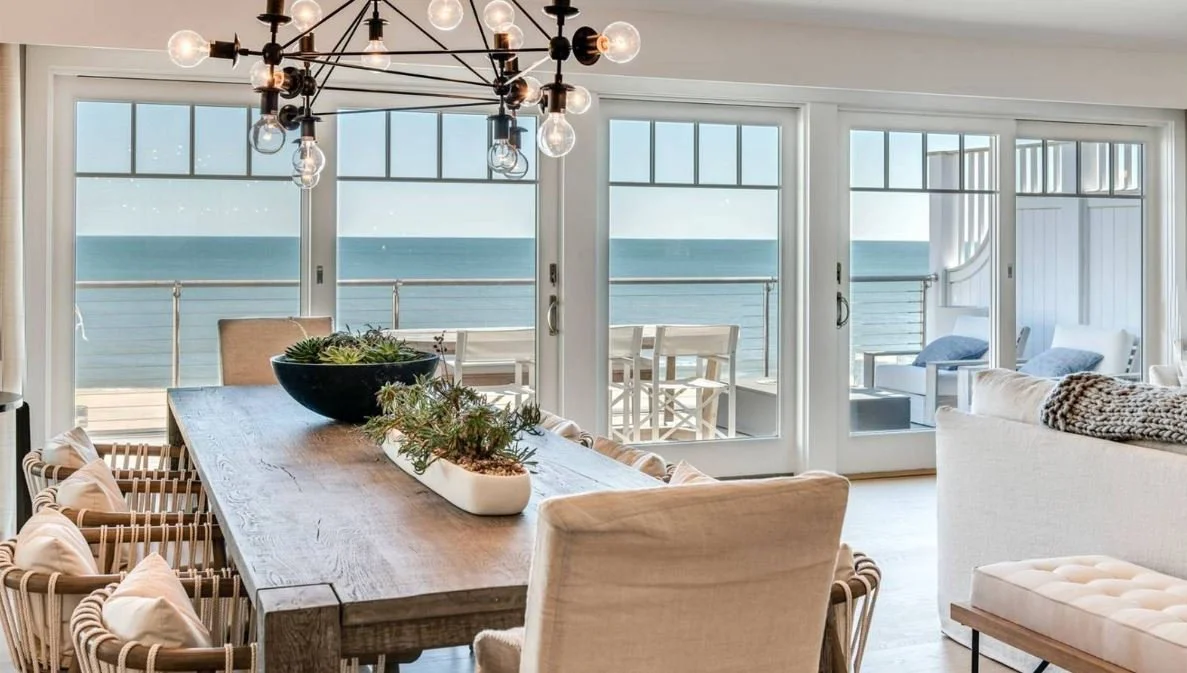 Oceanfront living room and terrace at the Gurney’s Residences in Montauk, featuring glass railings, pale wood outdoor seating and an unobstructed view across the beach to the Atlantic Ocean. Design