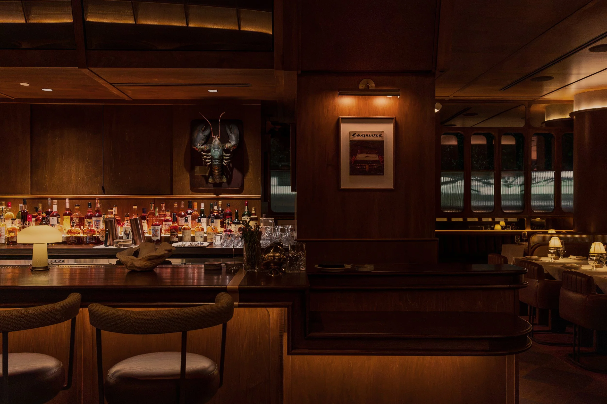 A framed artwork and sculptural wall light punctuate the richly detailed bar, where warm illumination highlights the bottles and dark millwork.