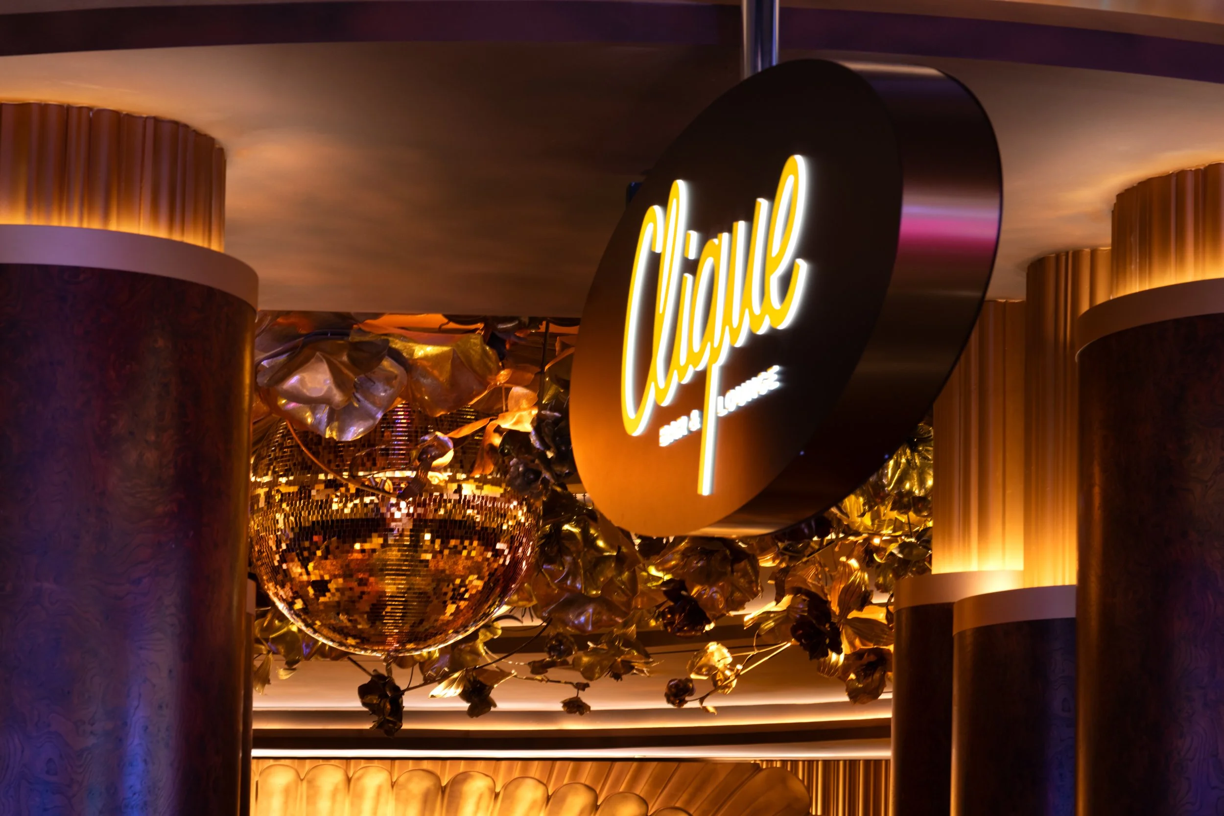 Clique Bar & Lounge at The Cosmopolitan of Las Vegas, featuring a glowing lounge sign, brass detailing, a disco ball, sculptural ceiling elements, and warm ambient lighting.