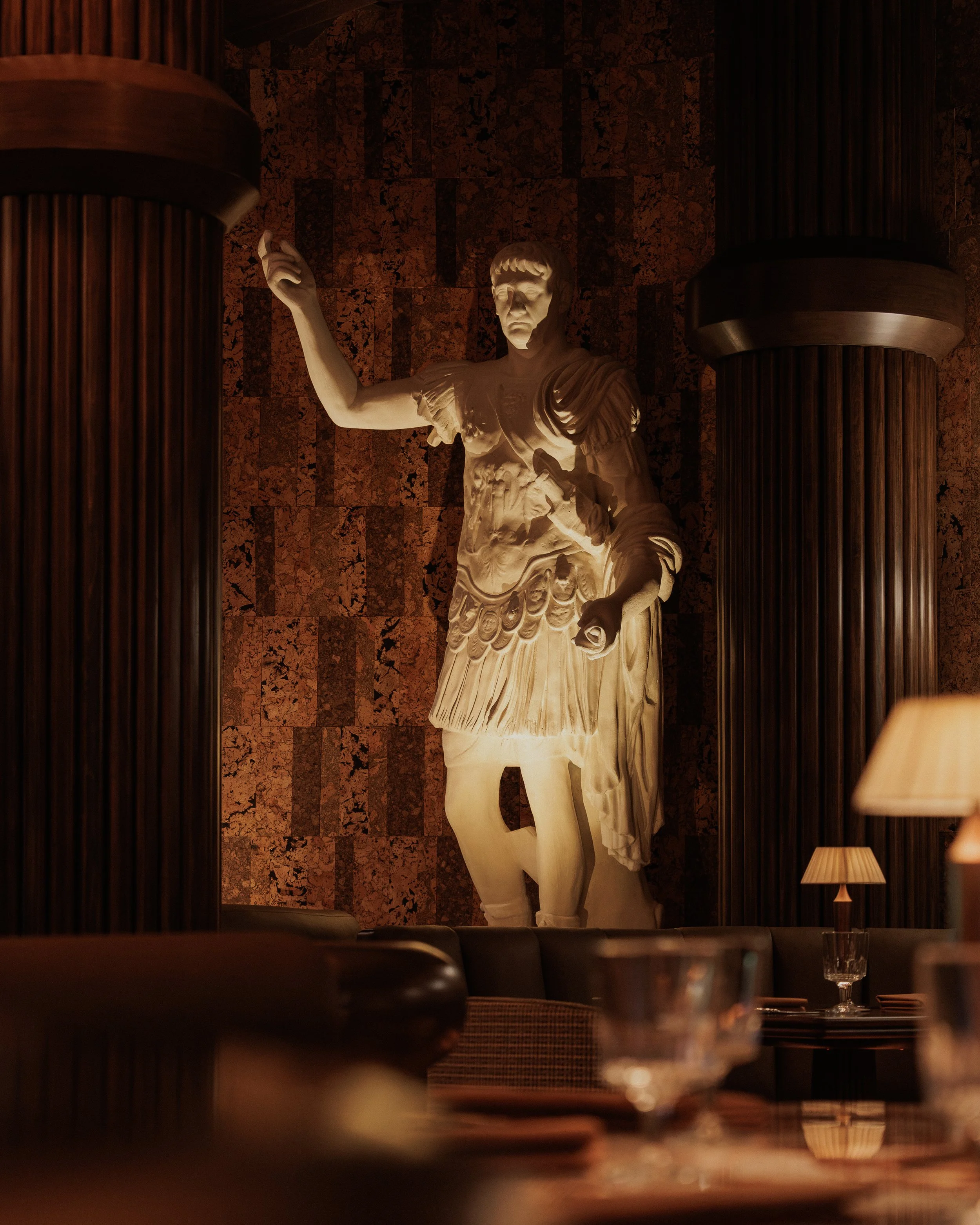 Illuminated Roman figure standing between dark fluted columns against a richly textured bronze wall in the dining room.