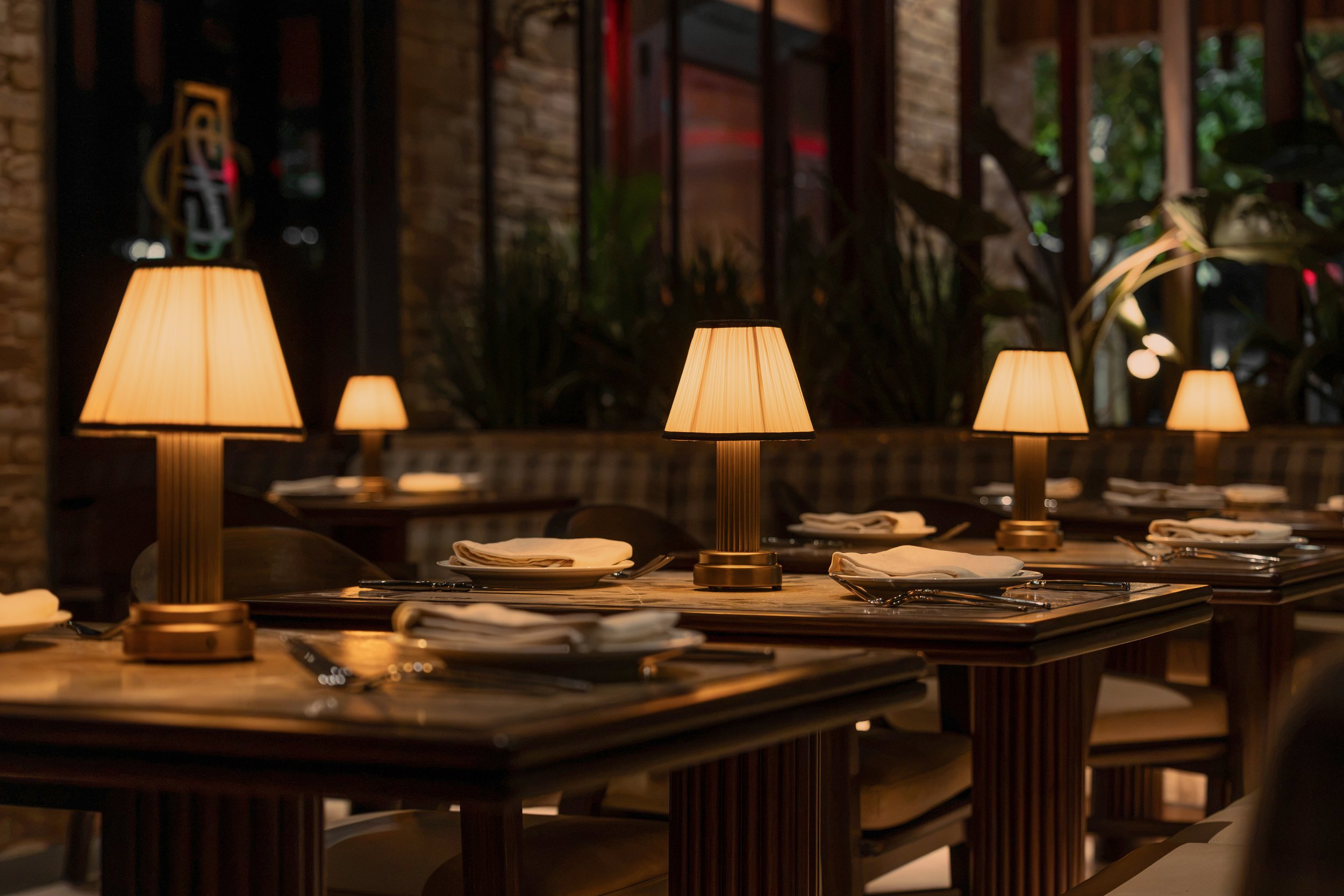 Close view of a dining table set with ceramic plates, glassware and shaded lamps against a backdrop of lush greenery.