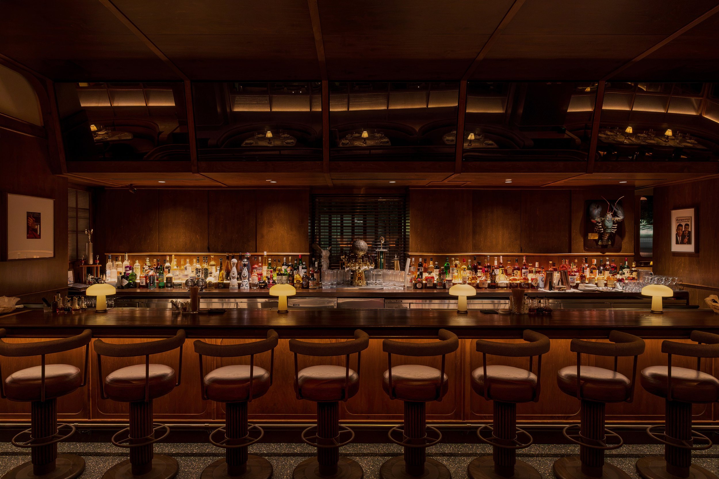 The central bar combines illuminated bottle displays, dark wood millwork and tailored counter seating beneath a reflective overhead canopy.