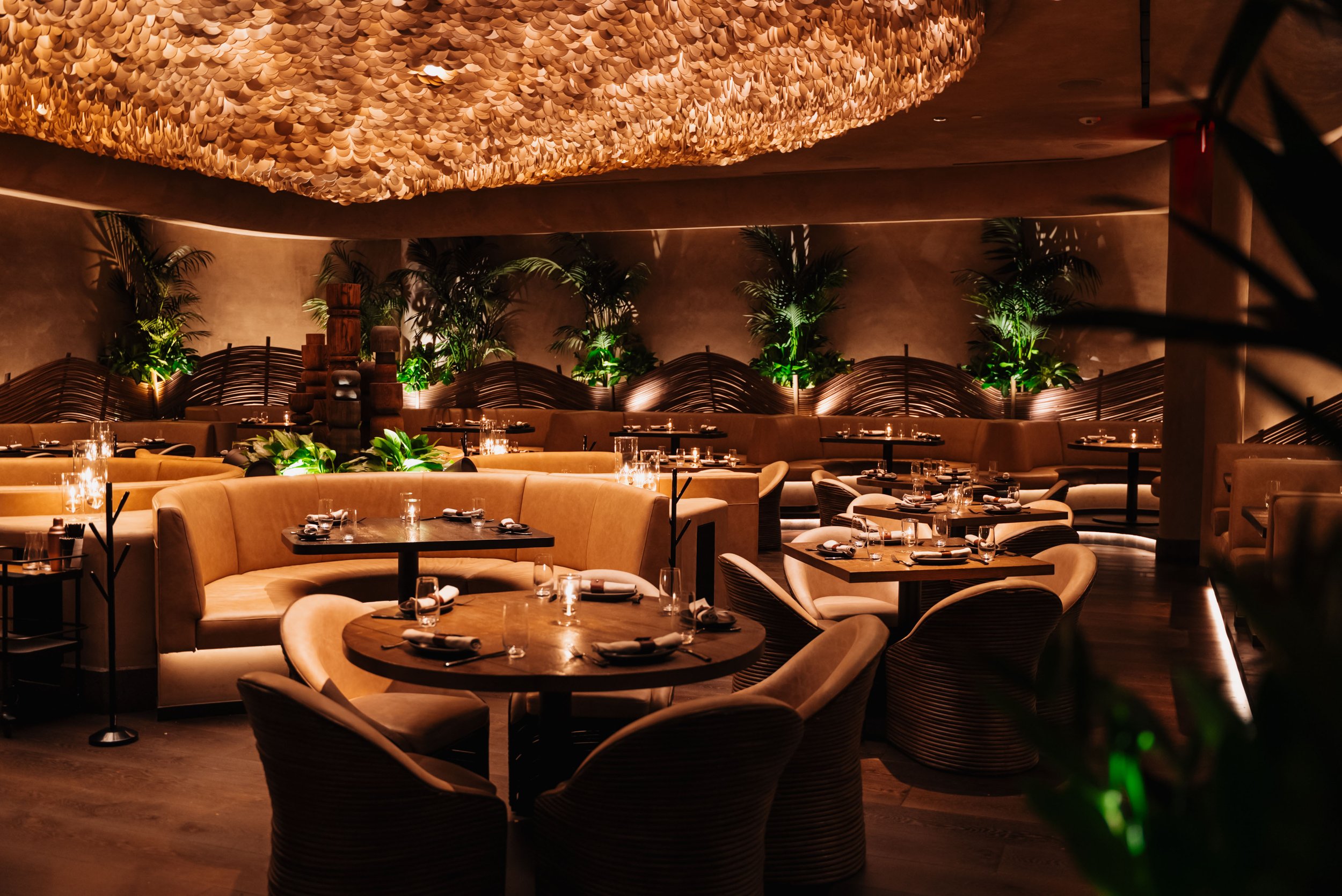 Dining room at Amaya inside The Cosmopolitan of Las Vegas, designed by PETERMAX Co. with intimate curved seating, warm amber lighting, dark wood and lush tropical planting.