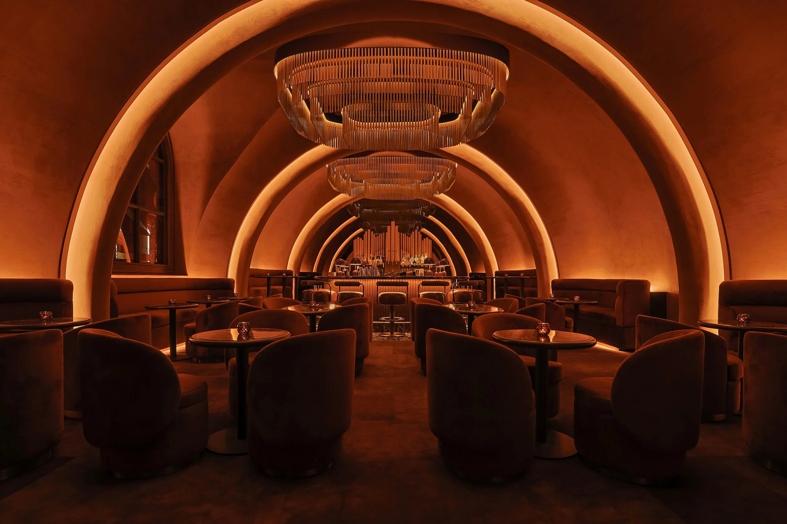 Long symmetrical view through Mea Culpa’s repeating illuminated arches and sculptural chandeliers toward the central bar.