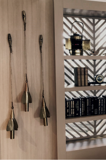 Decorative wall with hanging bells and a white wooden shelf filled with vintage books and decorative items, with a geometric-patterned window in the background.