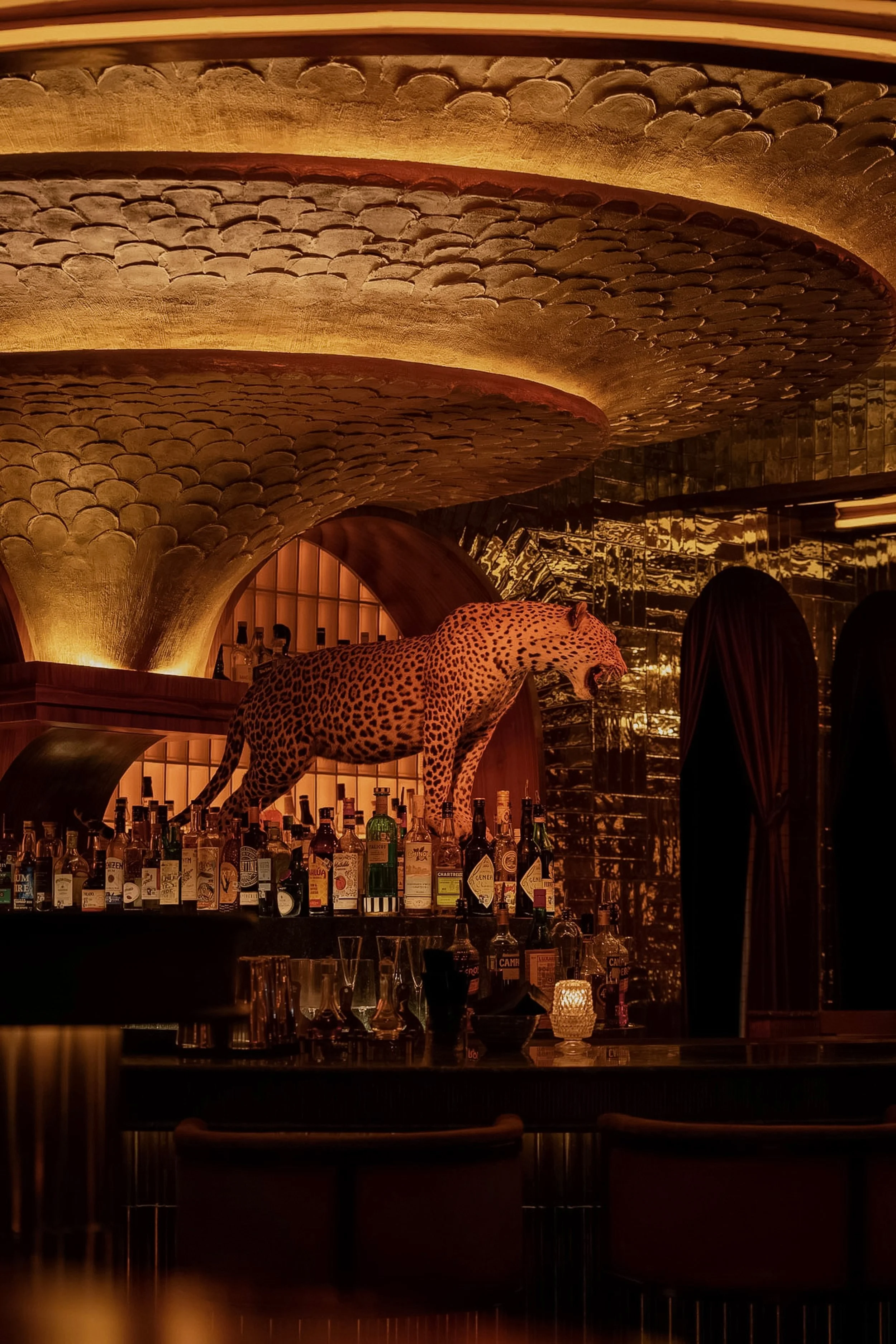 Powder Room’s central bar beneath a sculptural illuminated ceiling, with carved architectural details and a leopard figure overlooking the back bar.