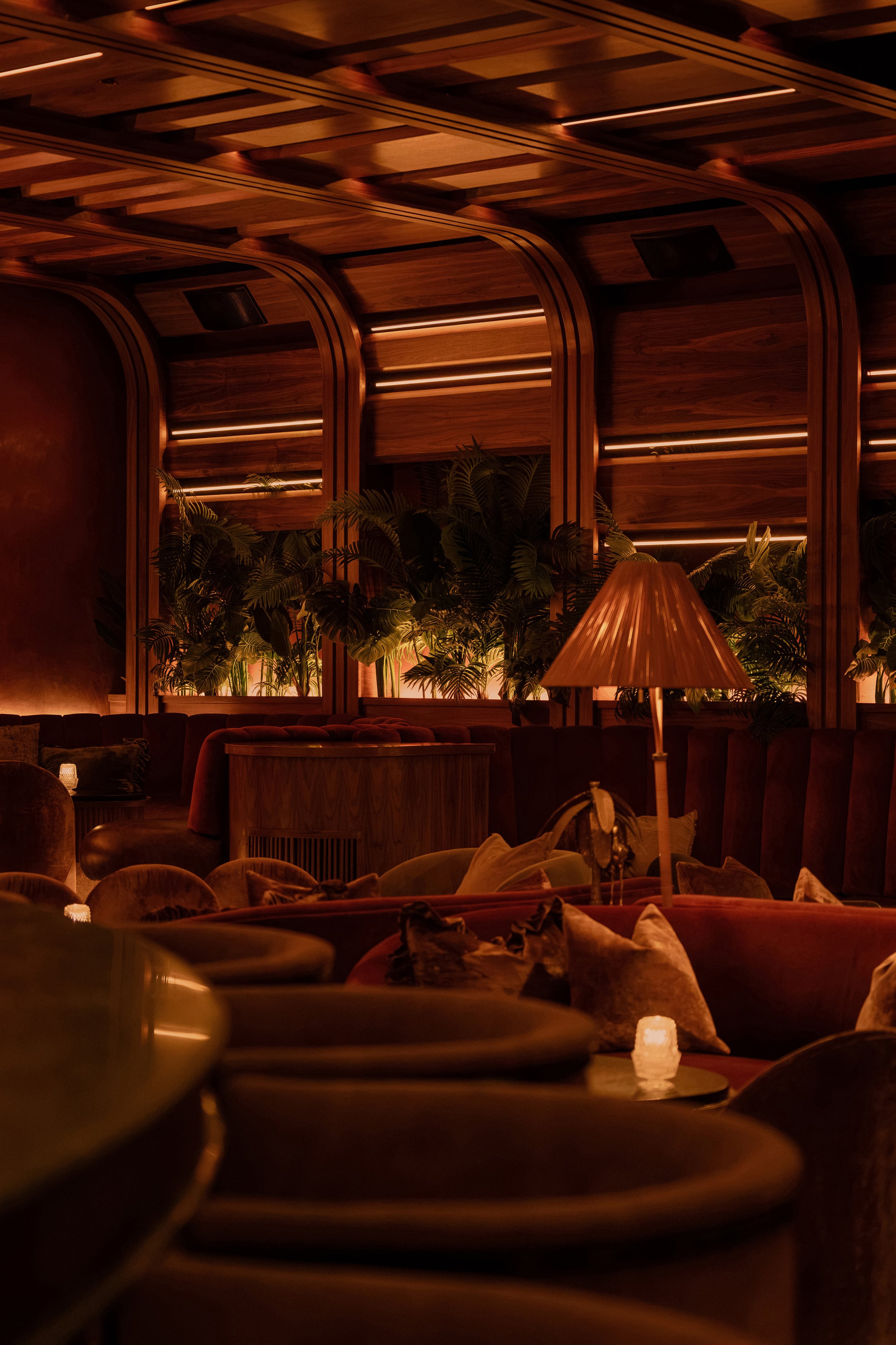 Layered lounge seating at Powder Room with velvet upholstery, tropical plants, wood paneling and a fringed table lamp.