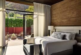 Hotel room with a bed, nightstand, and a sliding glass door leading to a balcony with outdoor seating and greenery.