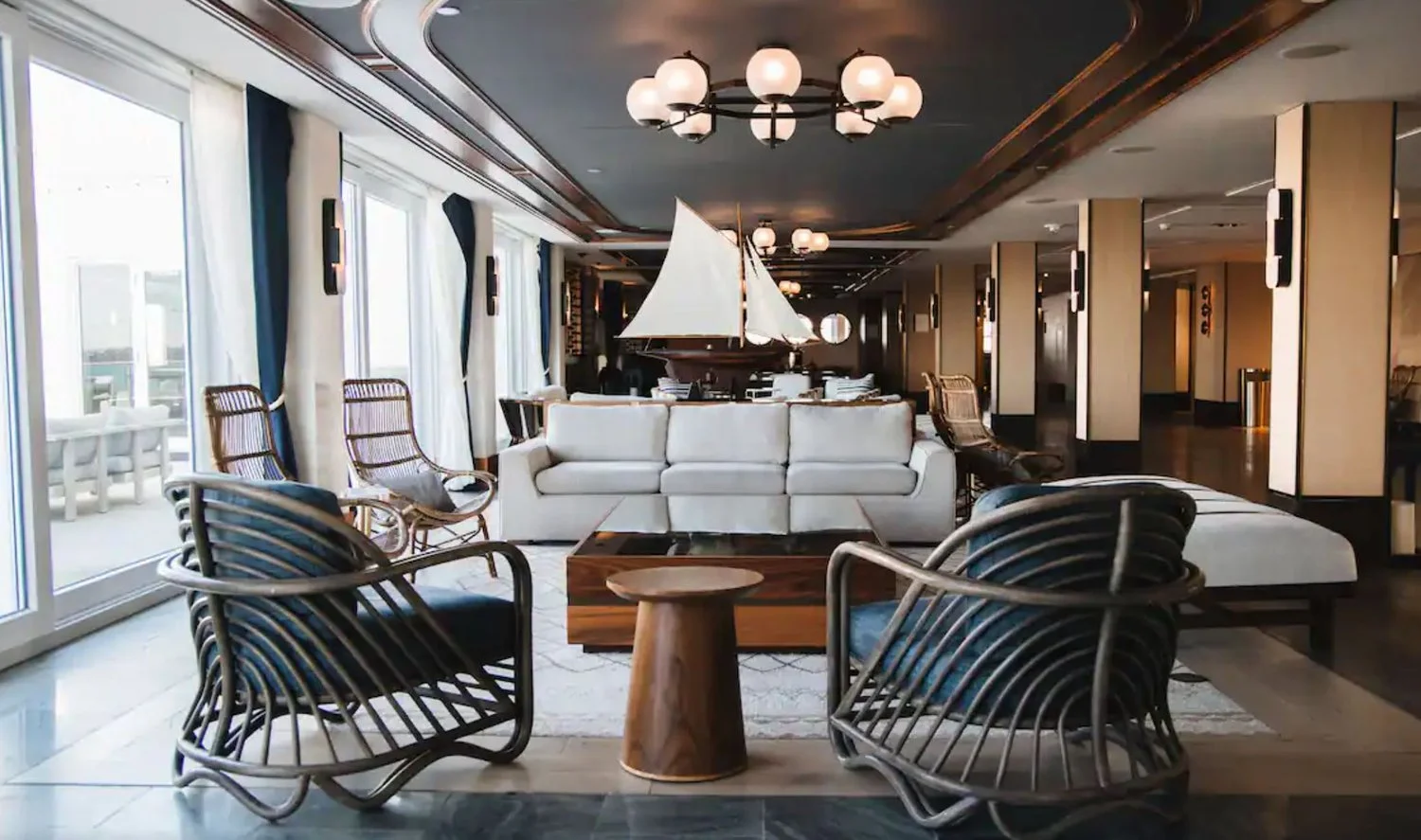 Elegant hotel lobby with large windows, modern chandelier, white sofa, wooden chairs, and nautical decor including a sailboat model