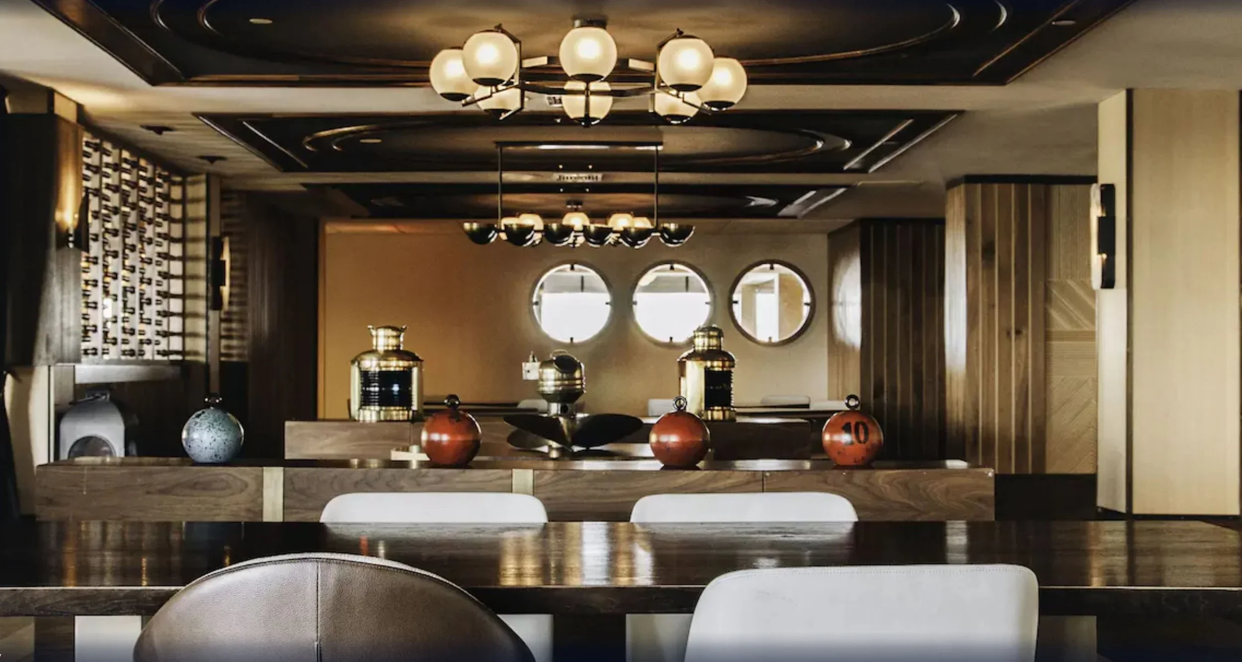Interior of a modern restaurant or cafe with dark wood furniture, decorative lighting including hanging and ceiling lights, and a wall with circular windows.