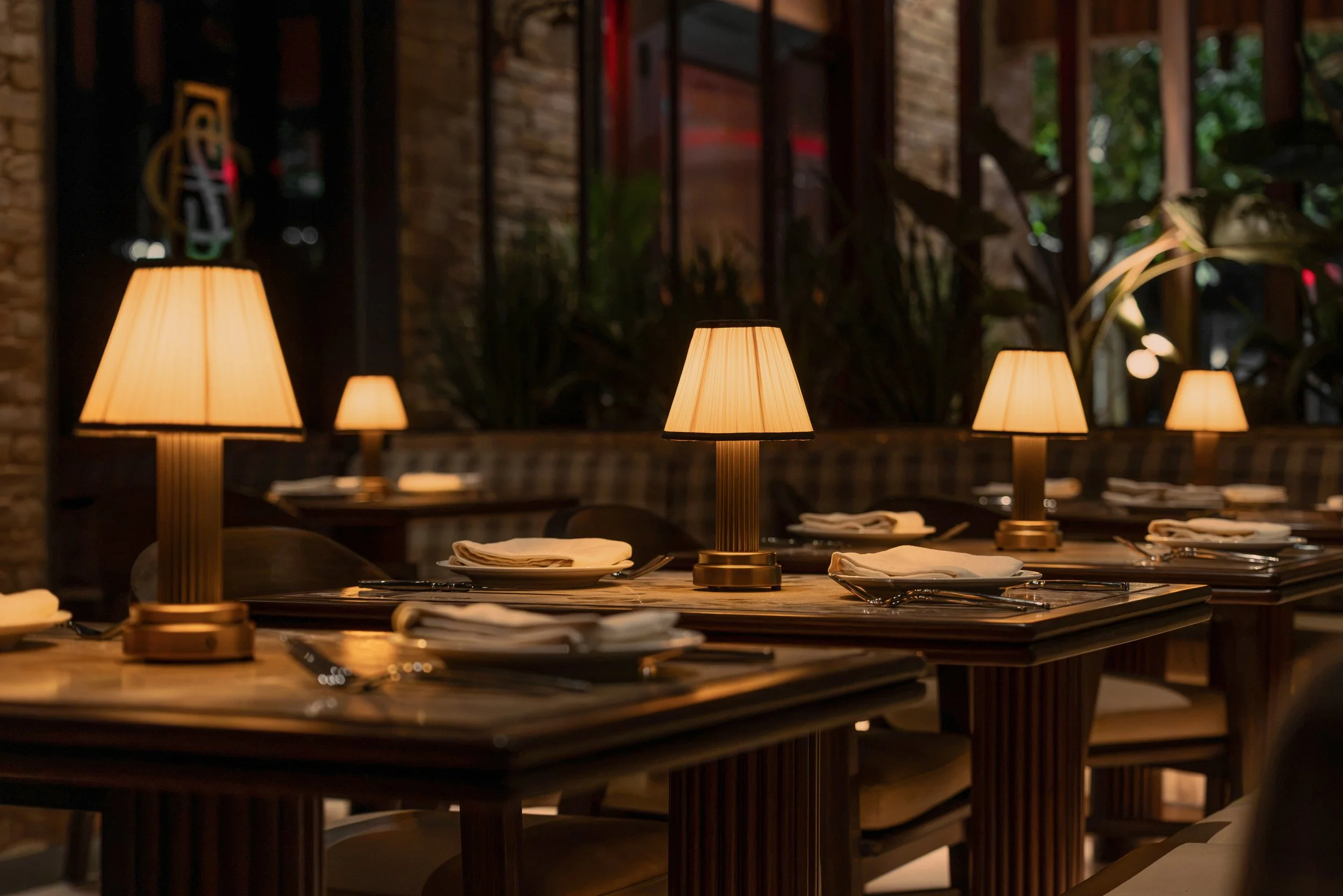 Close view of a dining table set with ceramic plates, glassware and shaded lamps against a backdrop of lush greenery.