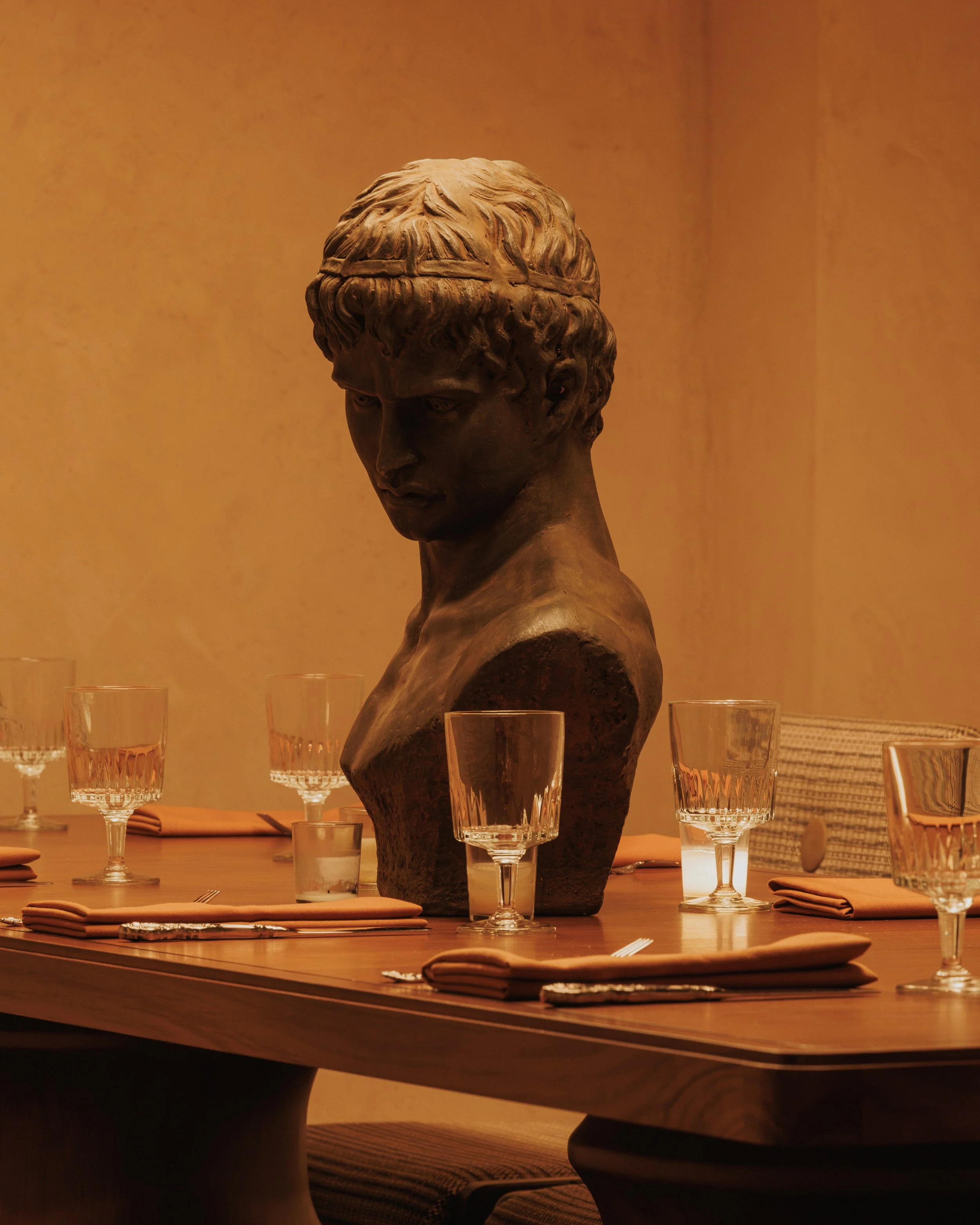 Classical male bust displayed beside a formally set dining table in the warm, atmospheric interior of Roman God of Fire.