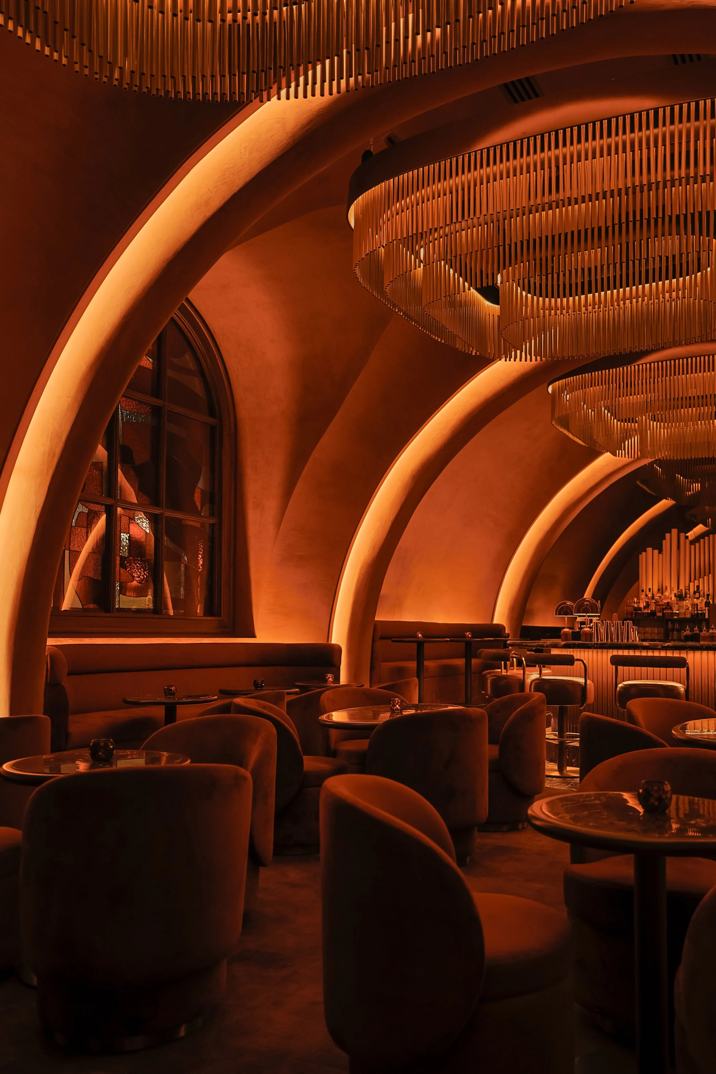Angled view across Mea Culpa’s marble bar toward curved banquettes, glowing architectural niches and sculptural metal chandeliers.