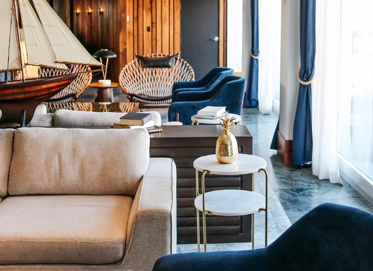 Luxury living room with beige sofa, small white marble side table with a gold pineapple decor, blue armchairs, a dining area with wicker chairs and model sailboats, wooden-paneled wall, and large windows with blue curtains.