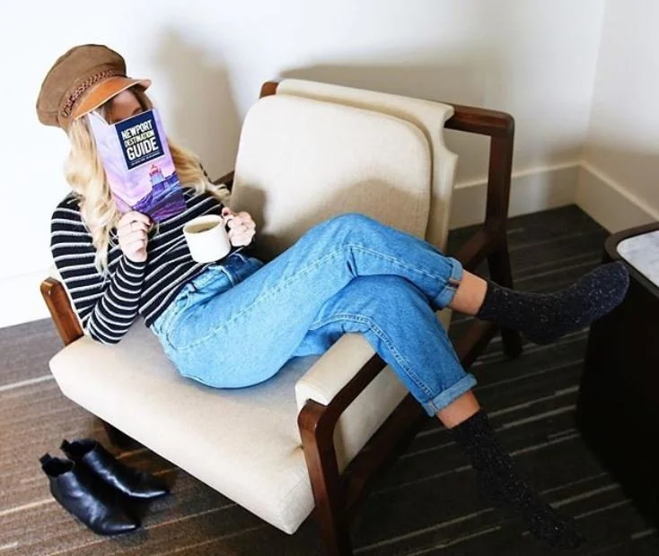 Woman sitting on a beige sofa, reading a guidebook, holding a coffee mug, wearing a striped shirt, jeans, a hat, and socks, with black shoes on the floor nearby.