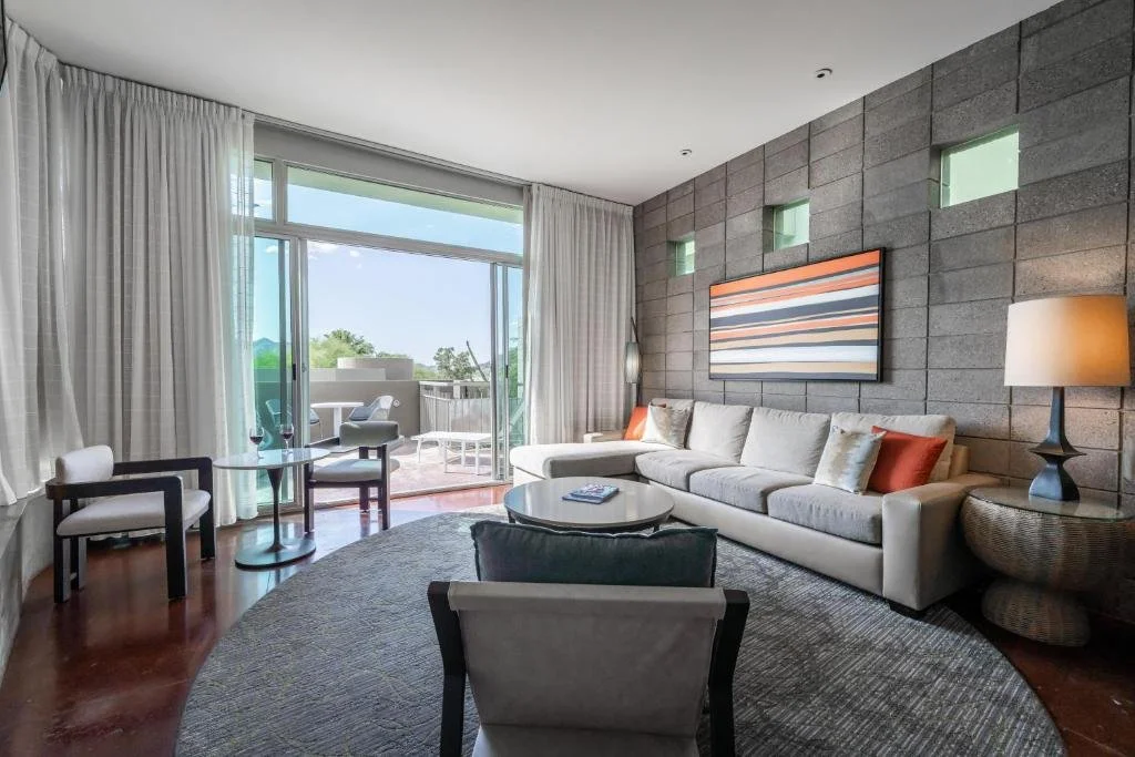 Living room with large gray couch, armchair, round coffee table, other chairs, and balcony with outdoor furniture, wall art, and textured gray wall.
