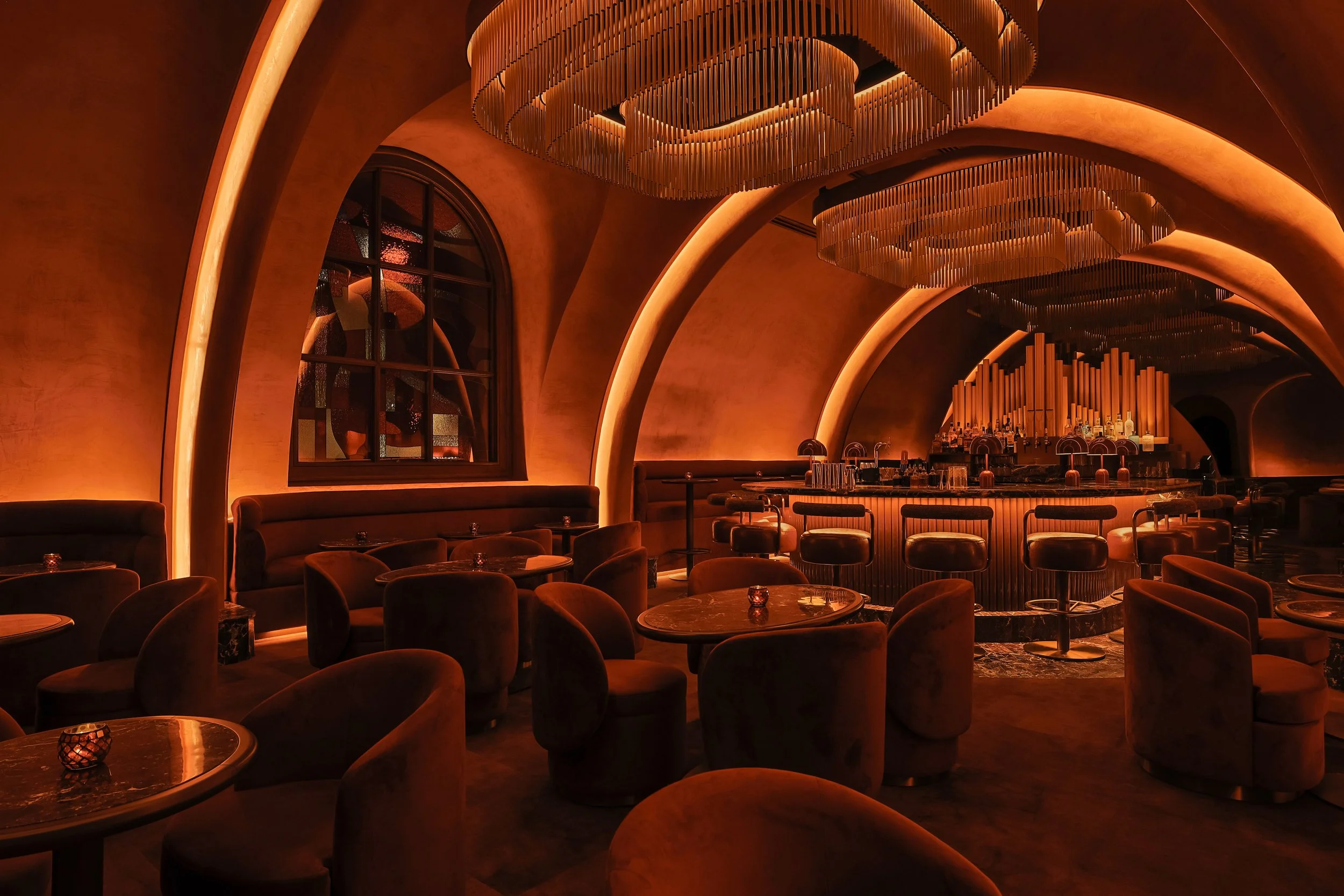 Angled view across Mea Culpa’s curved lounge seating toward the bar, with illuminated arches, sculptural chandeliers and an arched stained-glass window.