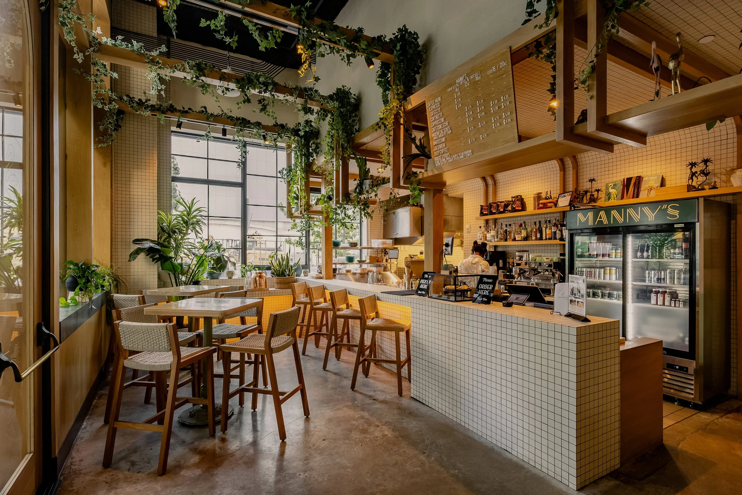 Sunlit interior of Manny’s featuring a tiled bar, wood stools, open shelving and cascading greenery.