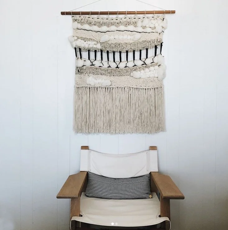 A wall hanging textile art piece with various textures and neutral colors hangs above a wooden chair with a black and white striped pillow.