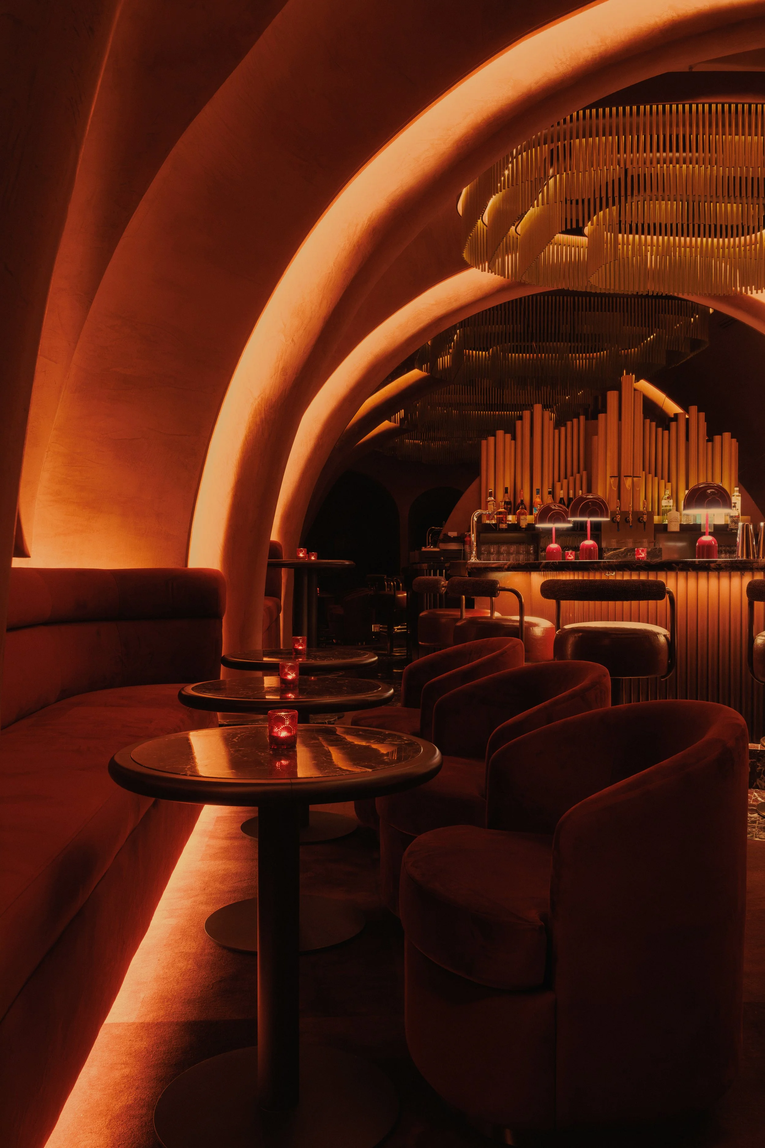 Curved marble bar at Mea Culpa with upholstered stools, red dome lamps, an arched display window and layered metal chandelier overhead.