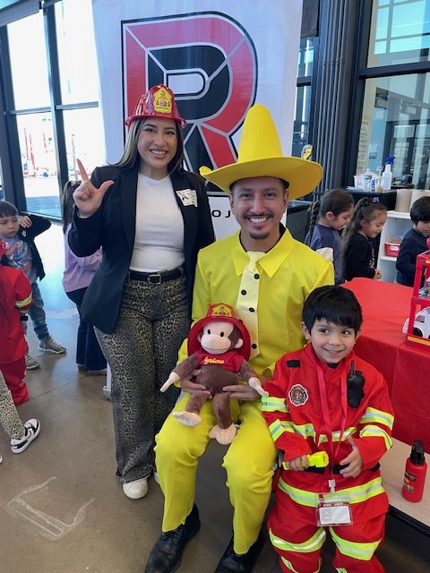 Group of diverse students in caps and gowns at a graduation ceremony, some reading, one smiling and making a peace sign, in a room with framed portraits on the wall.