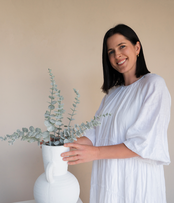 A woman with black hair, wearing a white dress, smiling while holding a white ceramic vase with eucalyptus branches inside.