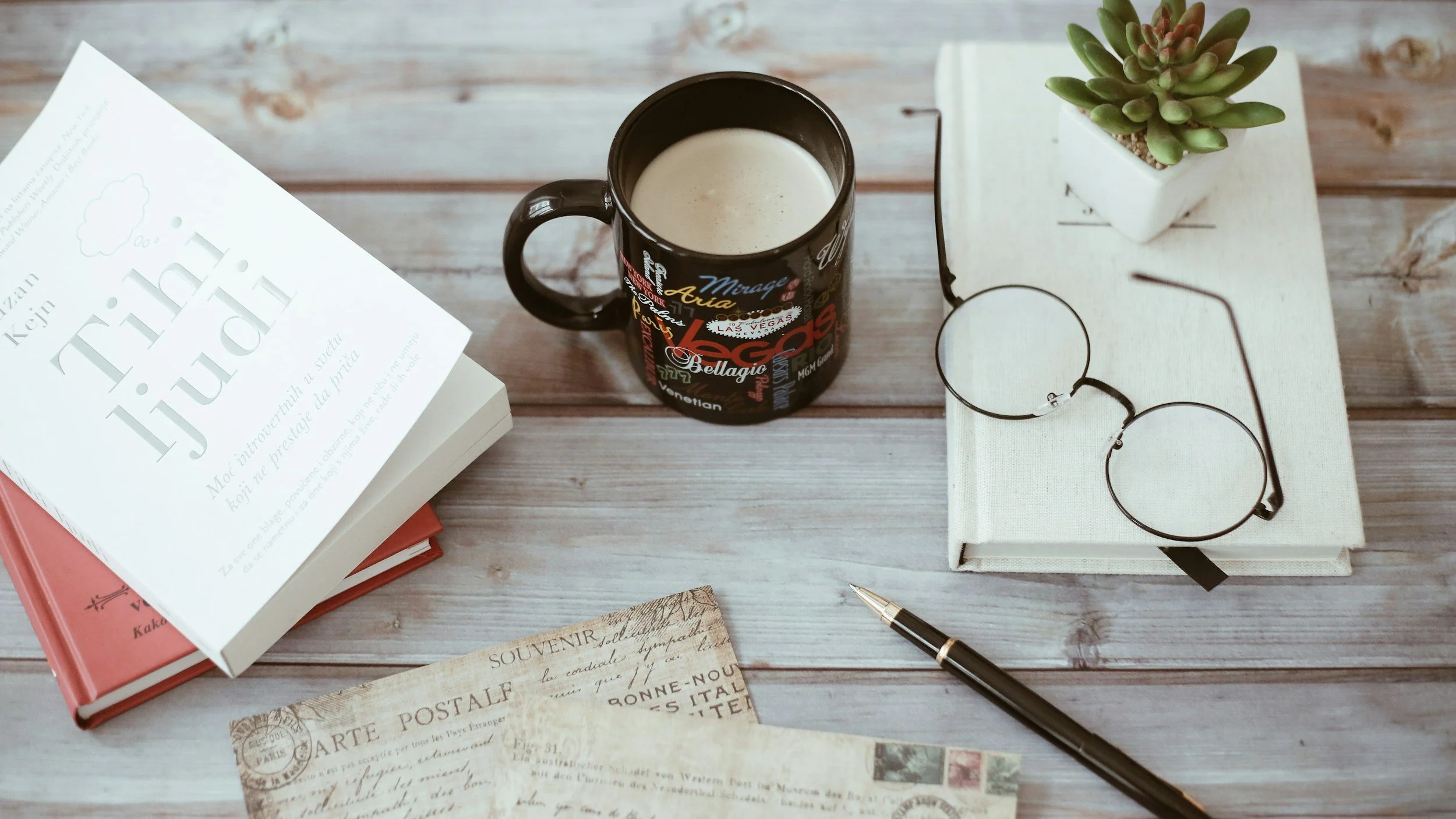A wooden table with books, a black coffee mug with coffee, a pair of round glasses, a pen, a small white plant pot with a succulent, and vintage postcards.