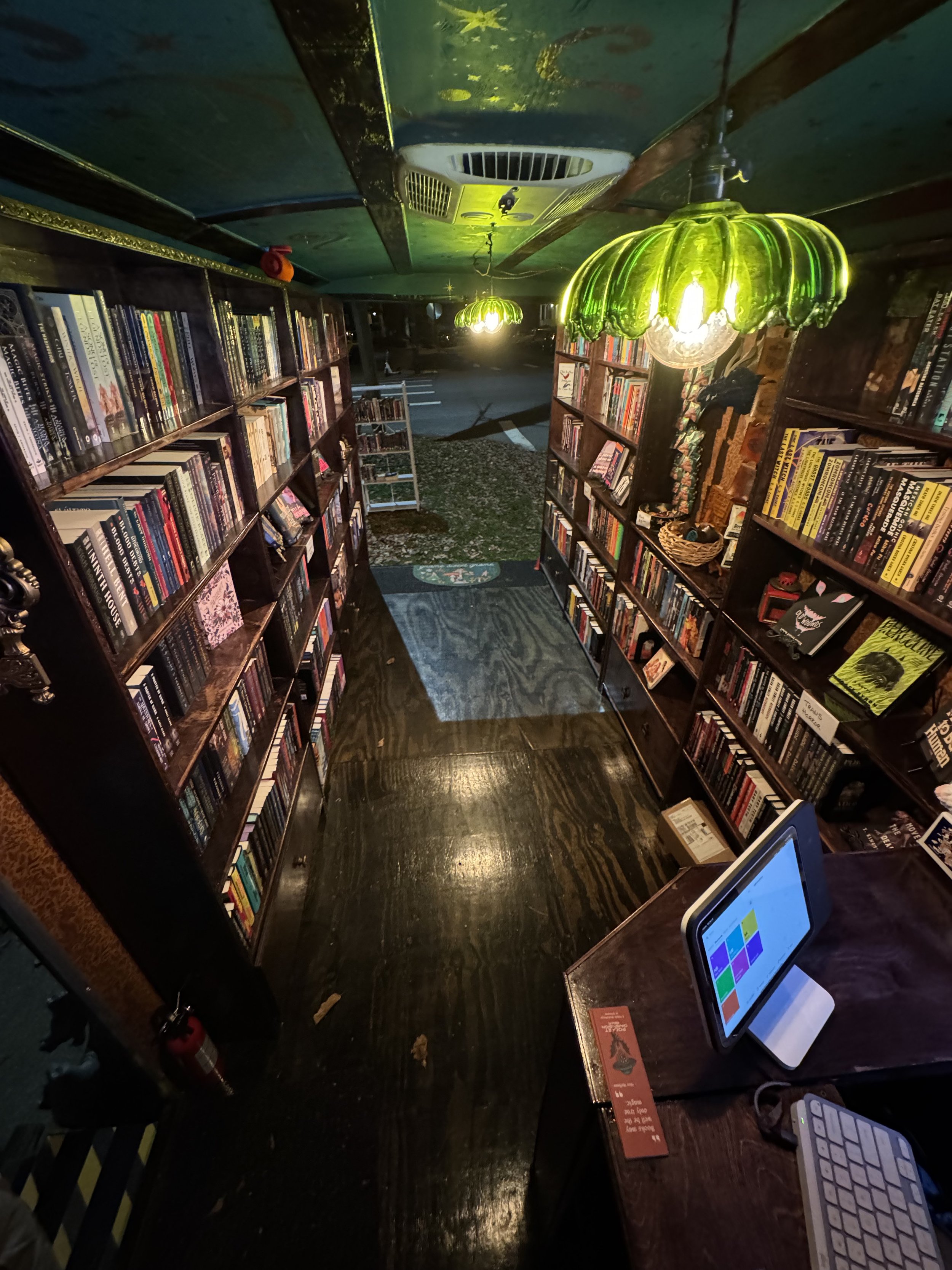 The inside of Pocket Dimension Bookshoppe at night. The green glass ceiling lights cast a magical glow on the wooden shelves lined with books. Painted metallic stars shine on the ceiling.