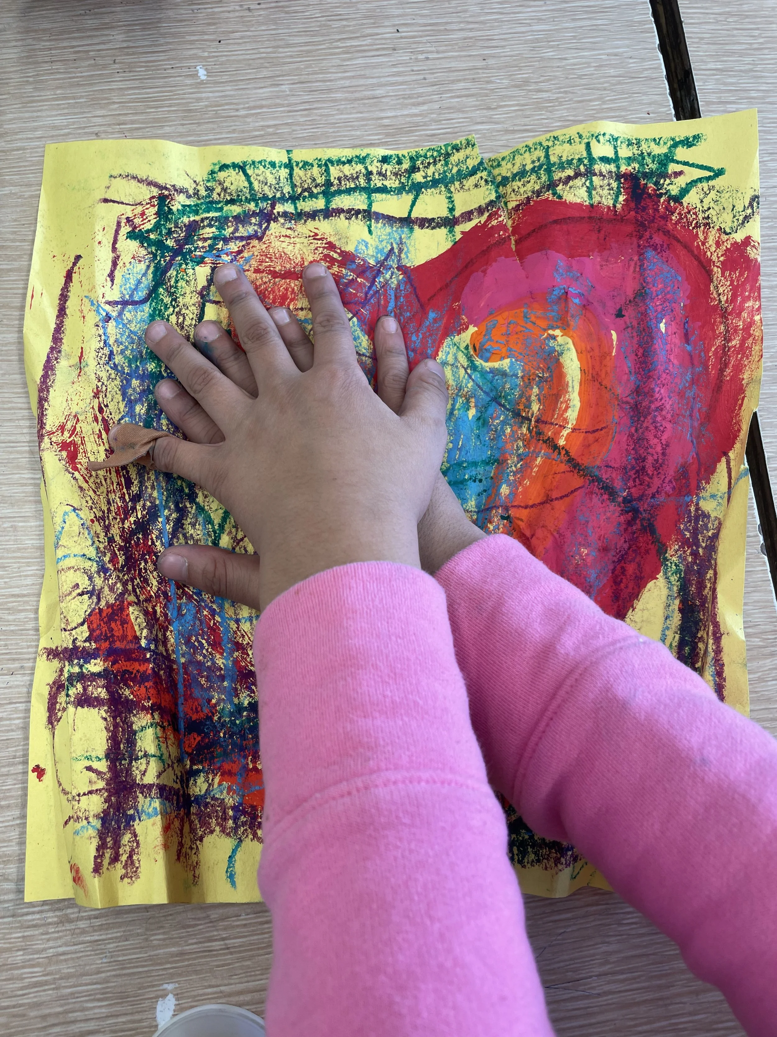 Child in pink sweater safely pressing hand onto a colorful abstract painting on yellow paper.
