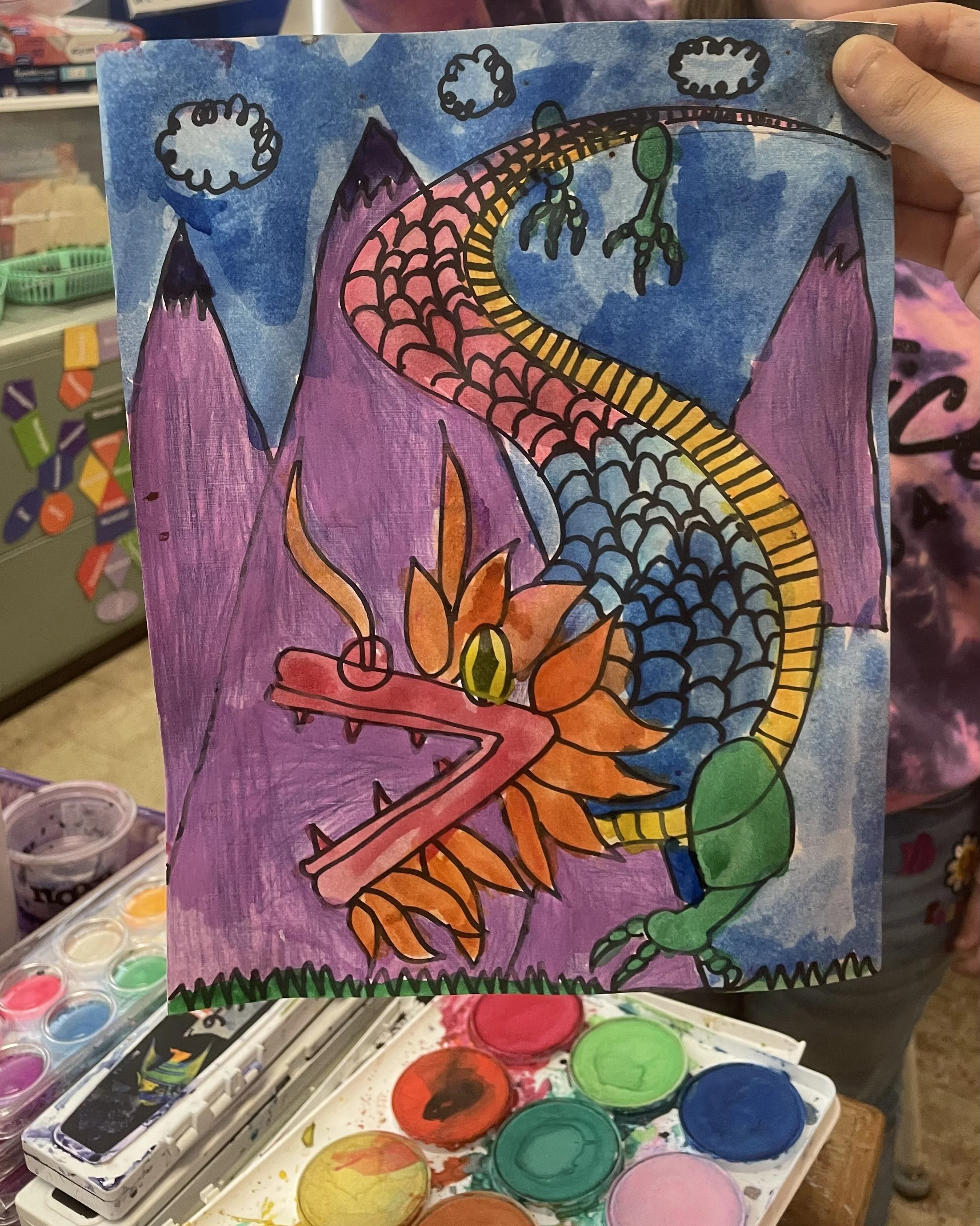 Children's colorful drawing of a dragon with purple mountains and a blue sky, created with watercolors and black outlining.