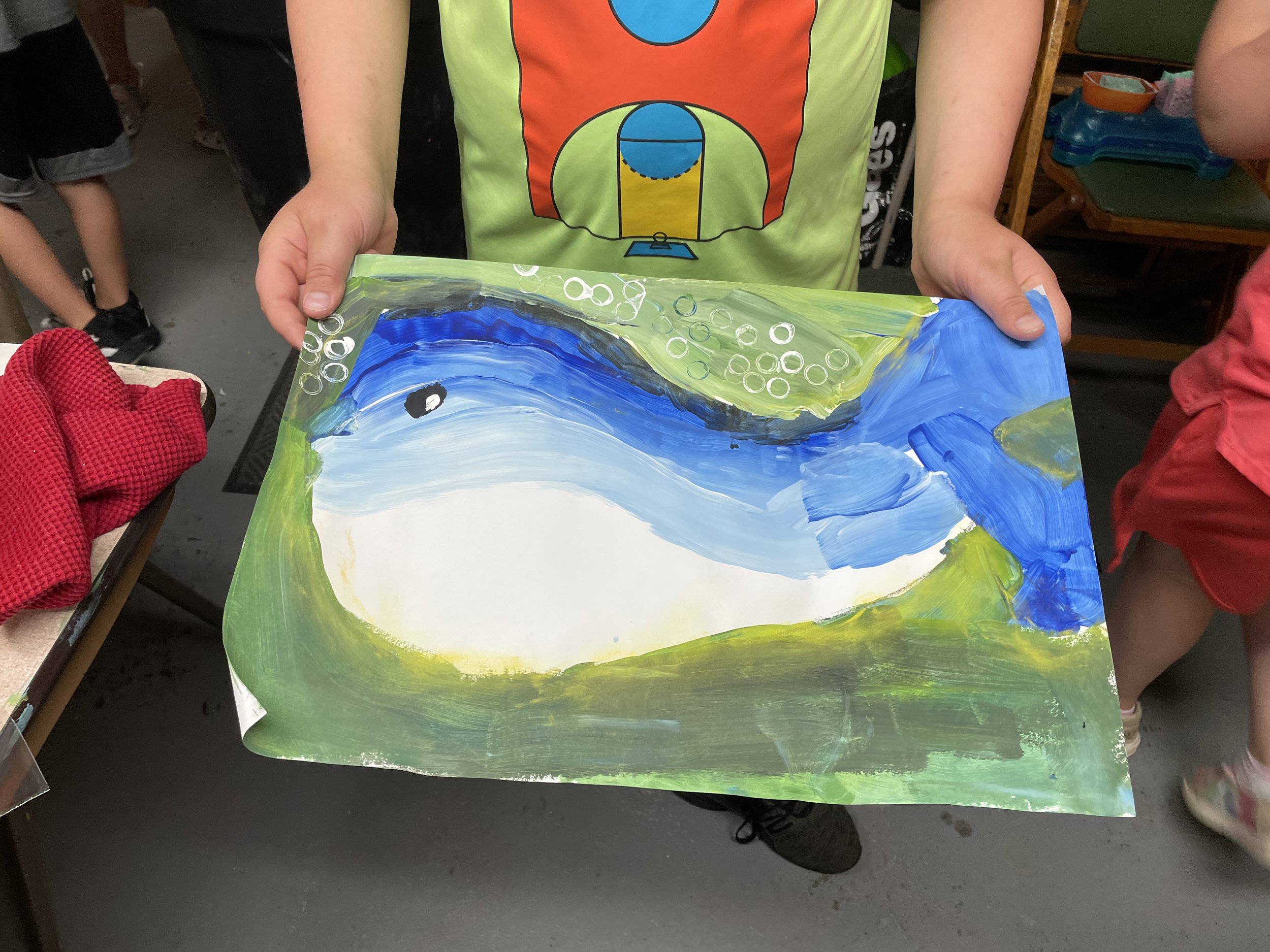 Child holding a painted picture of a blue whale on paper, with green, blue, and white colors, in a classroom setting.