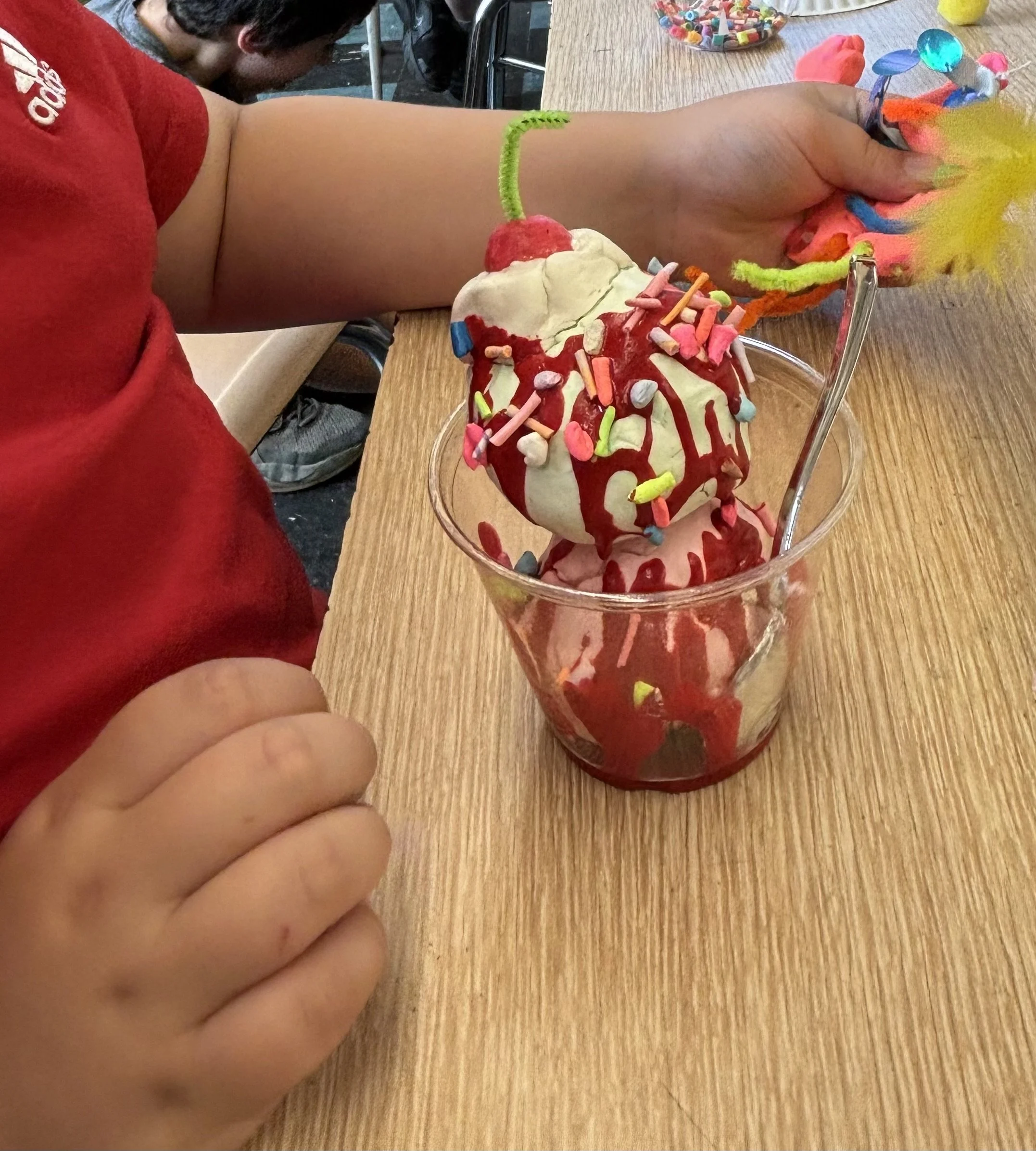 Child with a red shirt and tan skin sitting at a wooden table enjoying a colorful ice cream sundae with sprinkles, syrup, and a cherry on top, with another child blurred in the background.