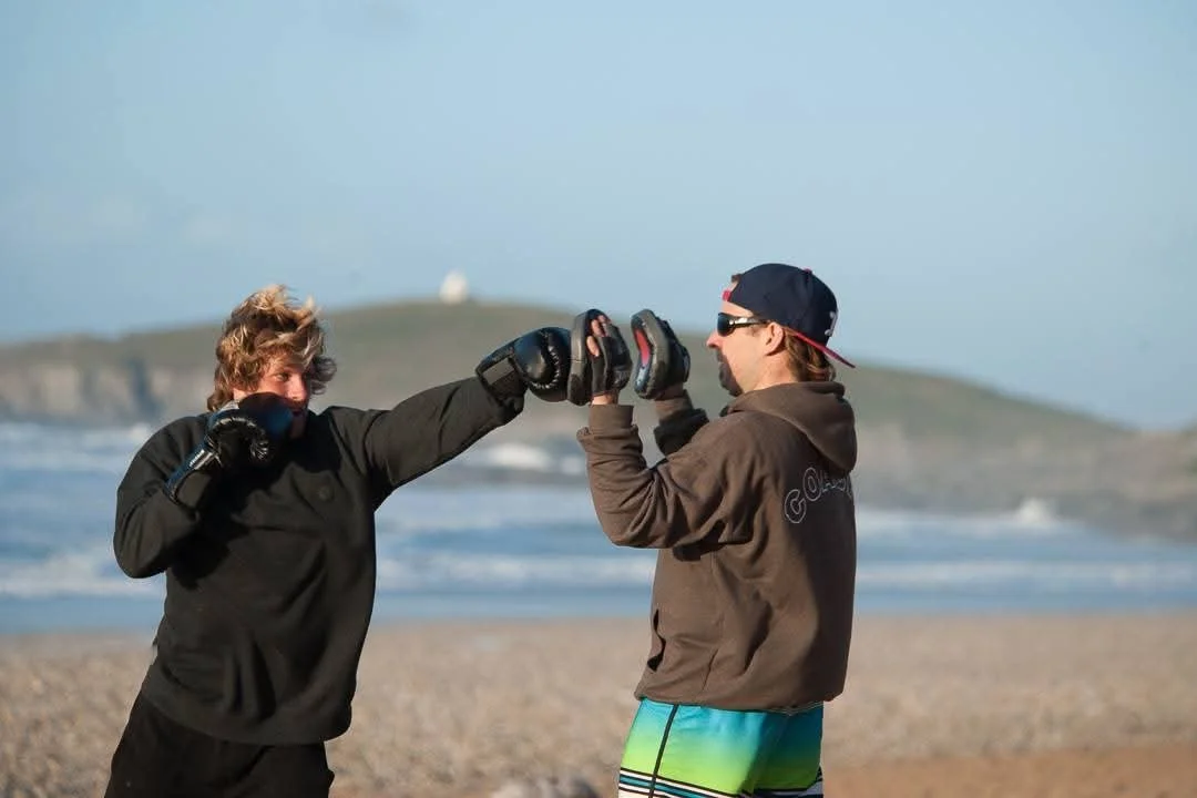A man getting a boxing lesson at the beach from another man wearing a hat and sunglasses.