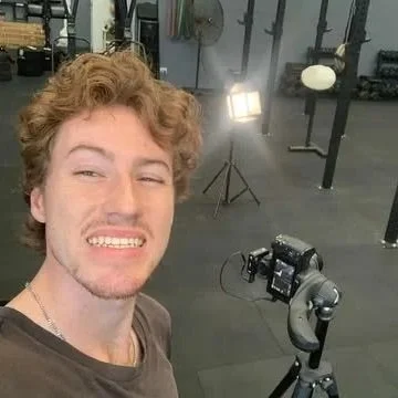 A young man taking a selfie in a gym, with a camera on a tripod behind him and gym equipment in the background.