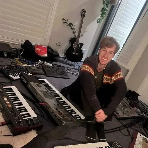 Young man sitting on the floor surrounded by music equipment, including synthesizers, a mixer, and a guitar in the background, smiling at the camera.