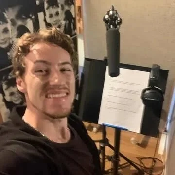 A man taking a selfie in a recording studio with a microphone and music stand in front of him.