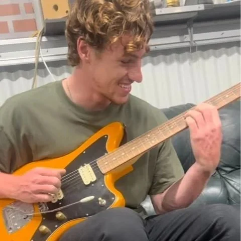 A man with curly hair playing an electric guitar, smiling, sitting on a couch in a room with colorful wall art in the background.