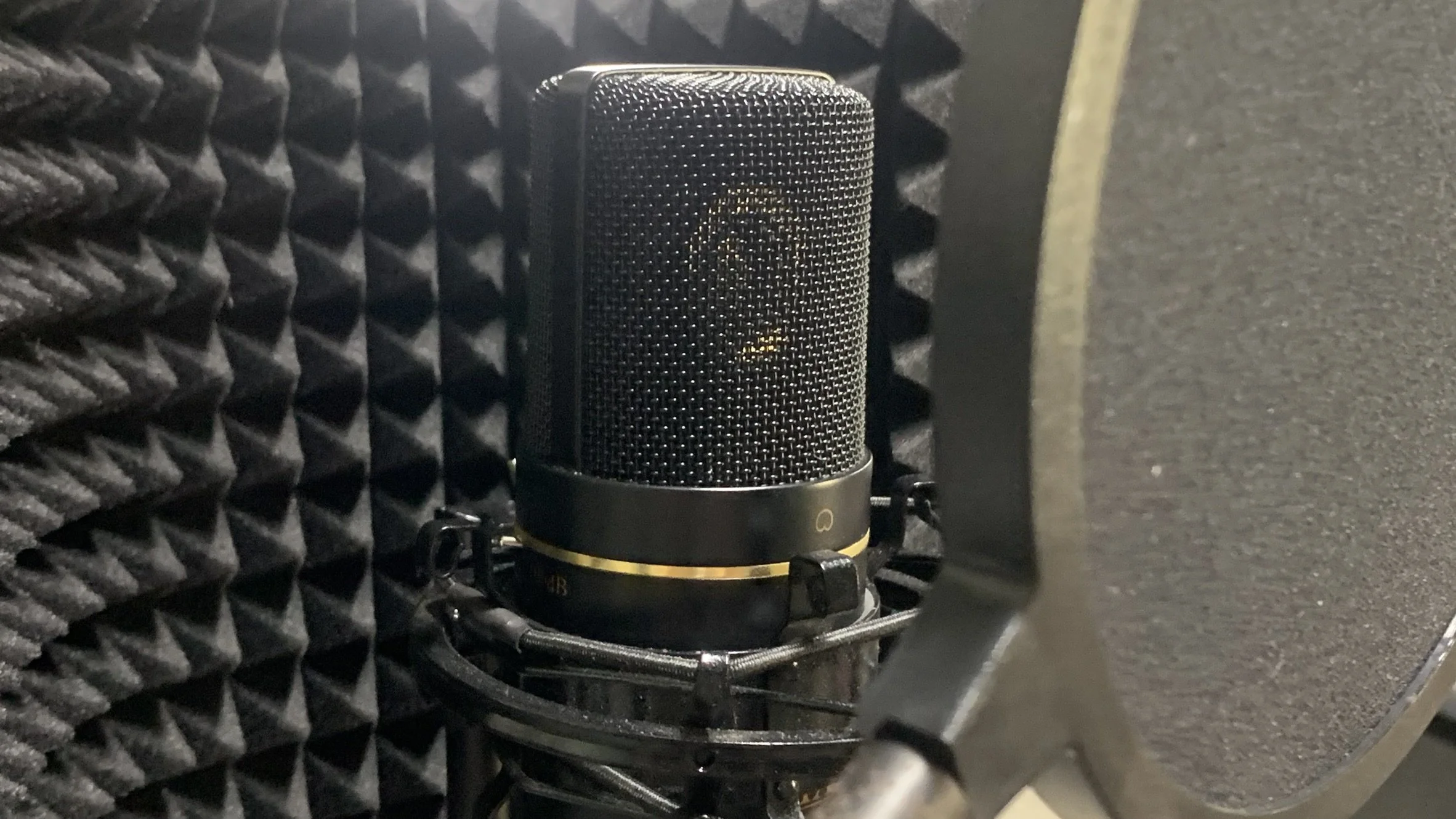 A condenser microphone with a metal mesh grille is mounted on a shock mount in a recording studio. The microphone is set in front of acoustic foam panels with pyramid-shaped soundproofing patterns, and a pop filter partially visible to the right.