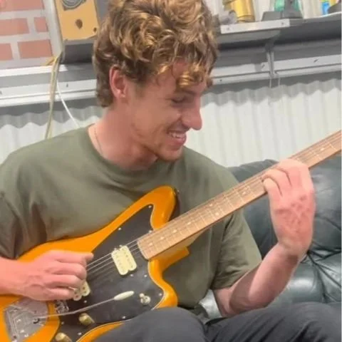 A young man with curly hair playing an electric guitar and smiling, sitting on a black leather couch in a room with shelves and boxes in the background.