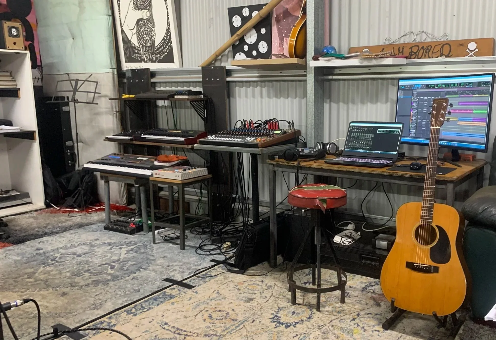 Music studio with various instruments and equipment including an acoustic guitar, keyboard, synthesizer, mixing console, headphones, computer, and speakers, all set up on tables and shelves with decorative artwork on the walls.