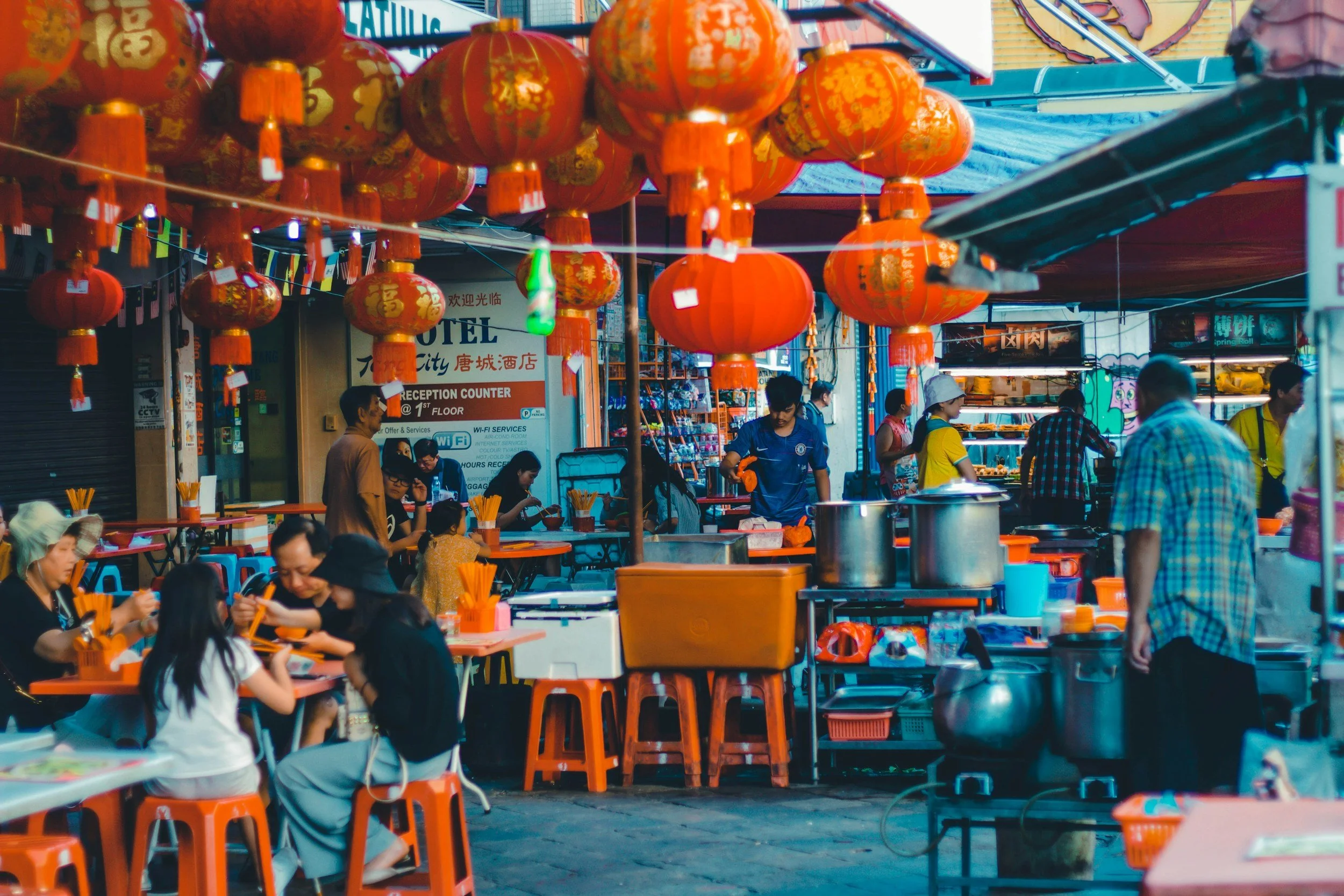 Street market with hanging red lanterns, people seated at orange tables, and vendors preparing food in an urban setting.