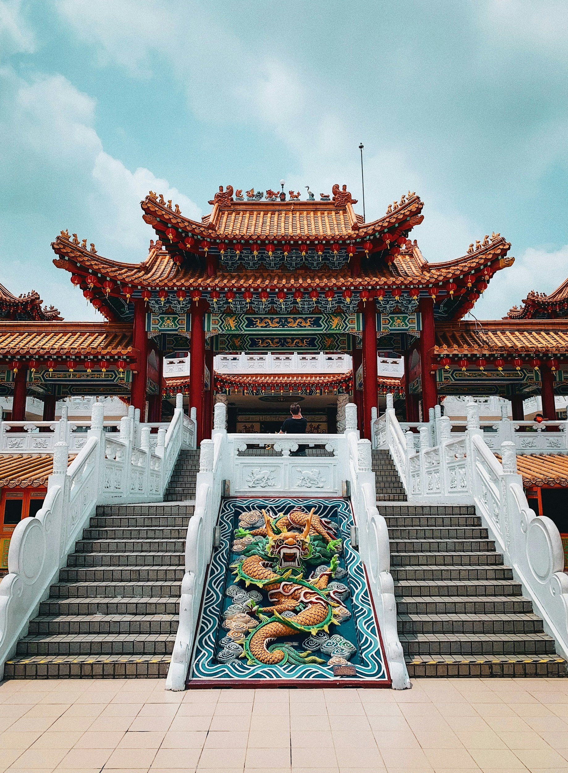 Traditional Chinese temple with stairs, colorful dragon sculpture at the base, ornate red and gold roof, and a person sitting on the upper platform.