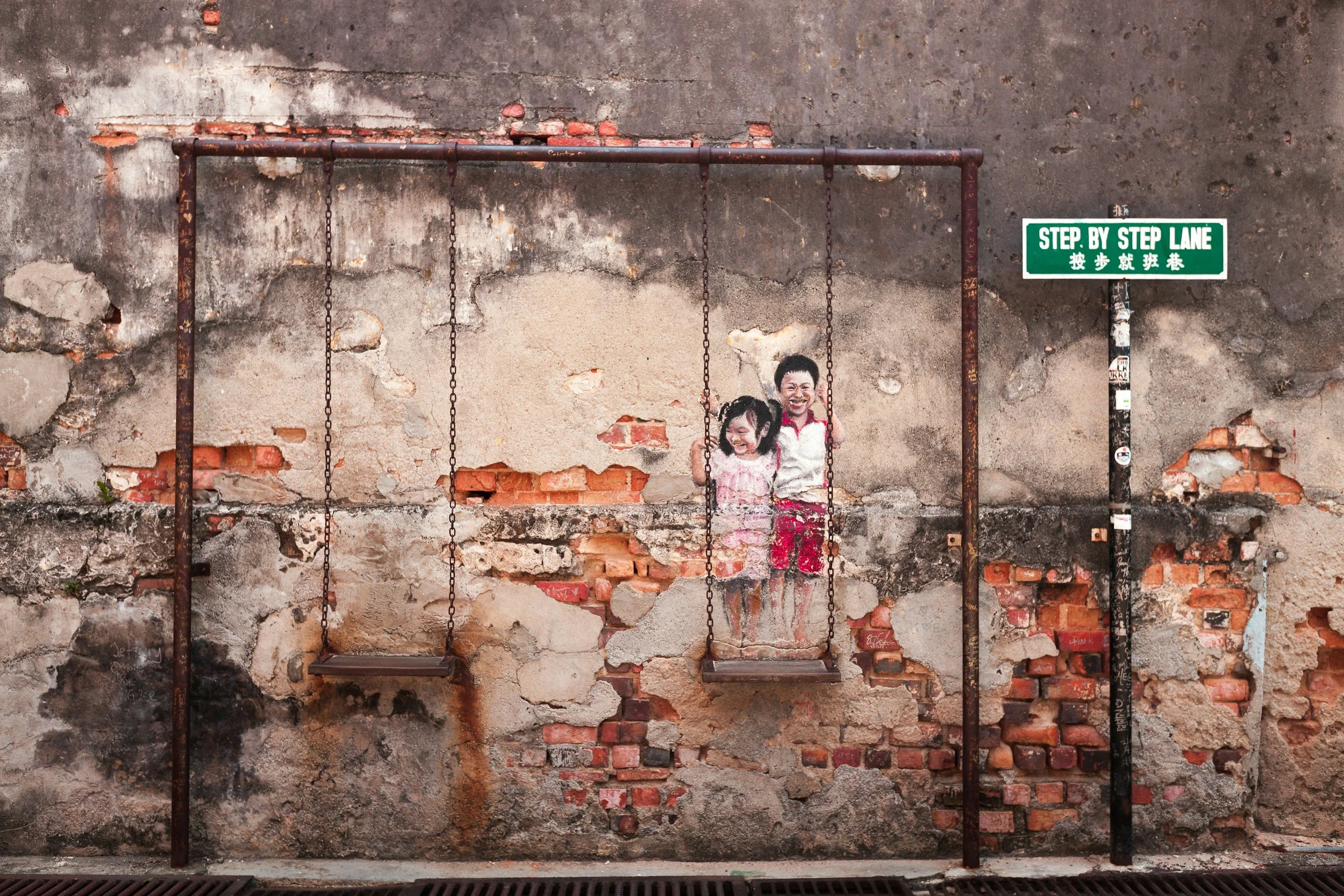 A mural of two smiling children on a rusty playground swing set against a worn brick and concrete wall, with a green street sign reading 'STEP BY STEP LANE' beside it.
